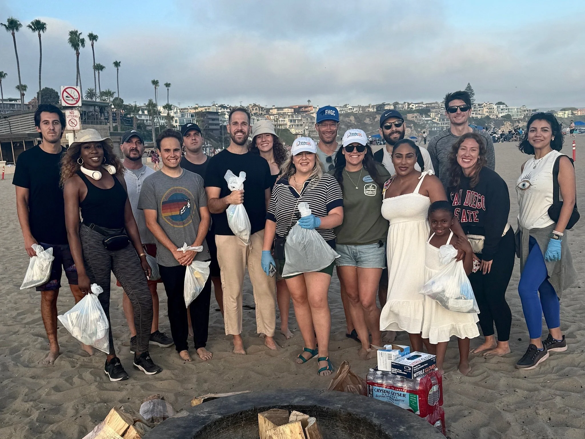Group of people on a beach holding trash bags during cleanup event, with houses and palm trees in the background.