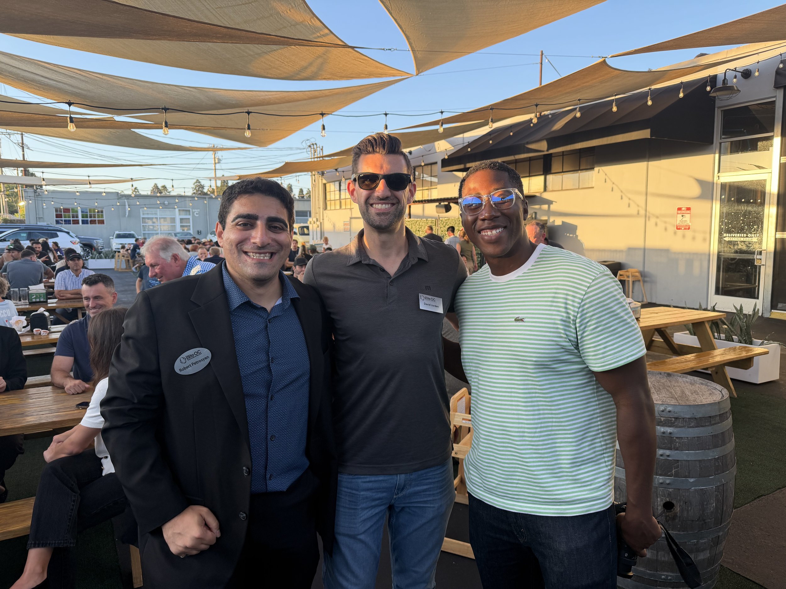 Three smiling men standing together at an outdoor event under shade sails, with other people sitting at tables behind them. The man on the left is wearing a dark suit with a name tag, the man in the middle is wearing sunglasses and a dark shirt, and the man on the right is wearing glasses and a green striped t-shirt.