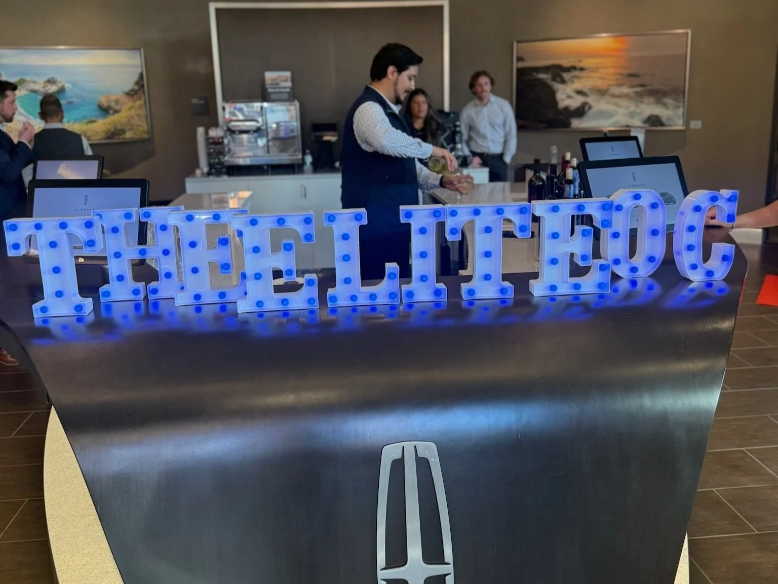 A reception area with a large illuminated sign spelling 'THE LOTUS.' A man is pouring a drink behind the counter, with several people present in the background, and art paintings on the wall.
