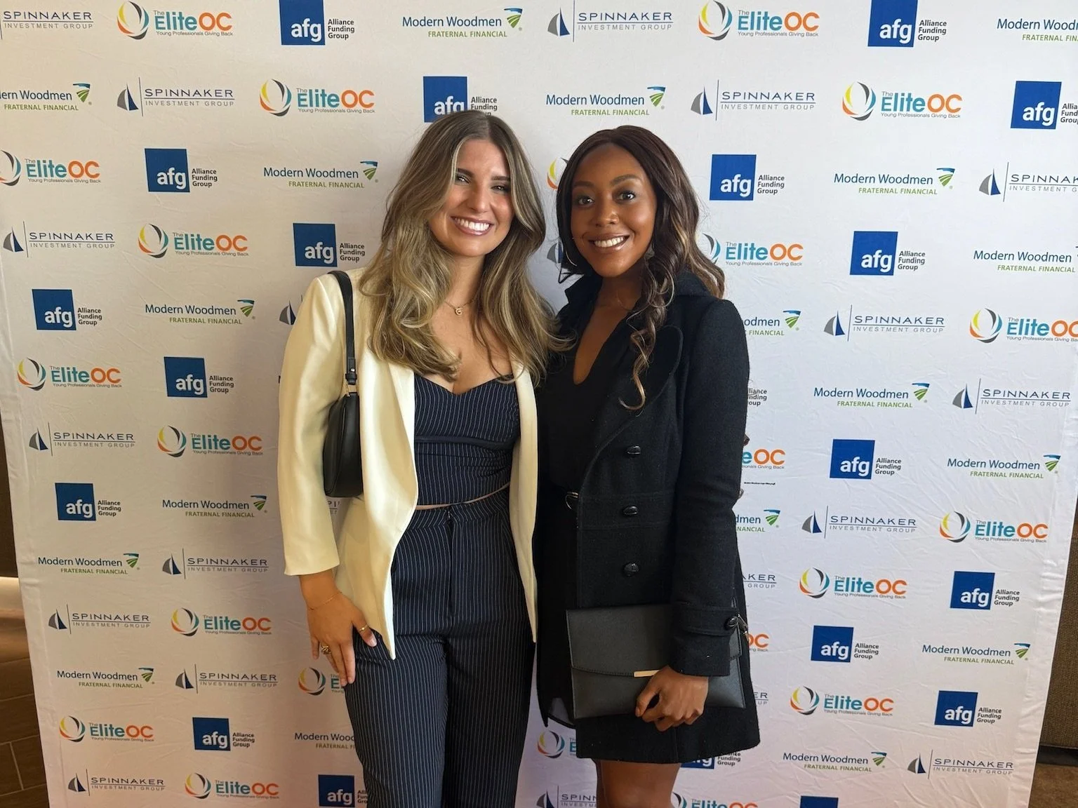 Two women smiling and posing in front of a backdrop with multiple logos, including Elite OC, aFG, Modern Woodmen, and Spinnaker Investment Group, at a professional event.
