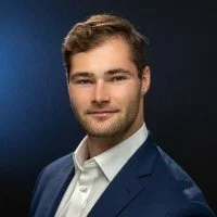 Portrait of a young man with short brown hair, wearing a dark blue suit and white shirt, against a dark background.