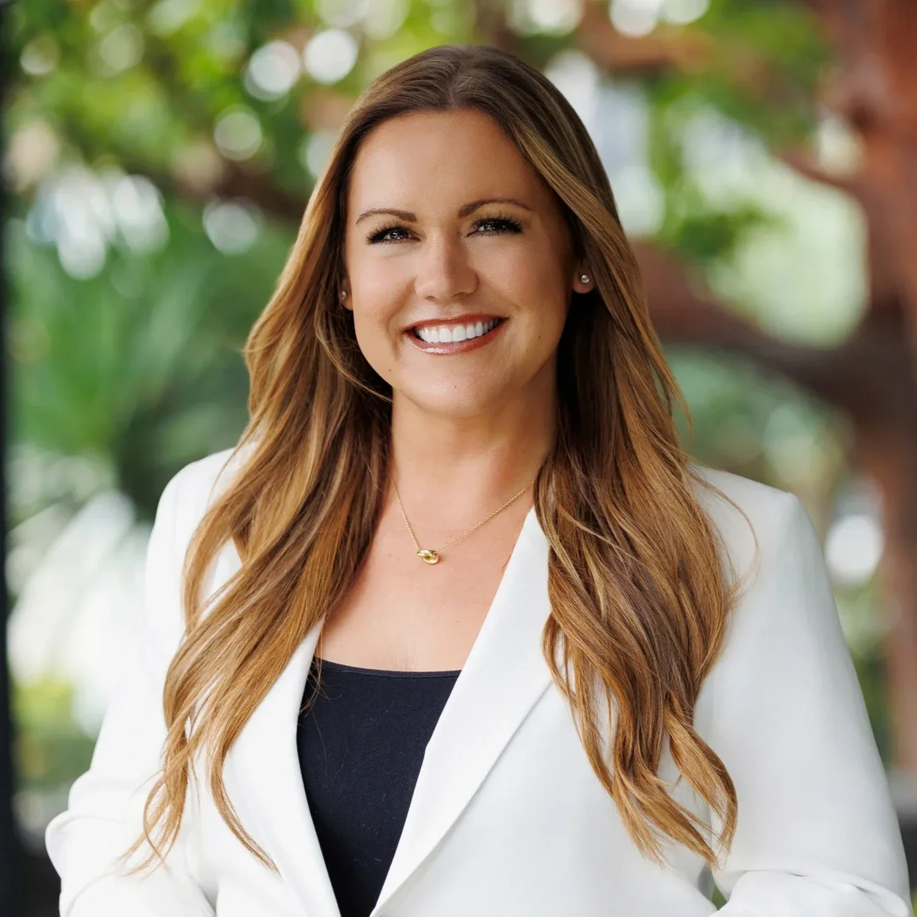 A woman with long, wavy, light brown hair smiling outdoors with green blurred foliage in the background, wearing a white blazer, a black top, and a gold necklace.