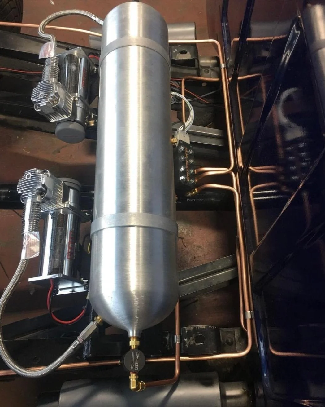 A close-up of a compressed air or gas system with a large cylindrical stainless steel tank, copper pipes, electrical components, and fittings mounted on a black metal frame.