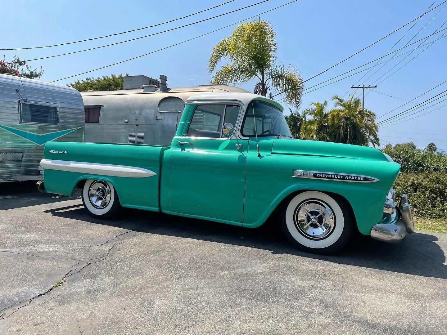 A vintage green Chevrolet Apache pickup truck parked on asphalt with a silver camper trailer behind it, against a backdrop of trees, palm trees, and power lines on a sunny day.