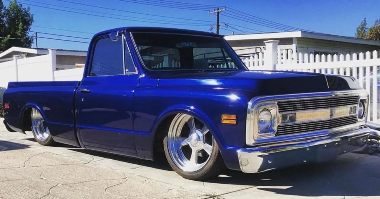 A vintage blue pickup truck with lowered stance and custom wheels parked on a driveway, with a white fence and clear blue sky in the background.