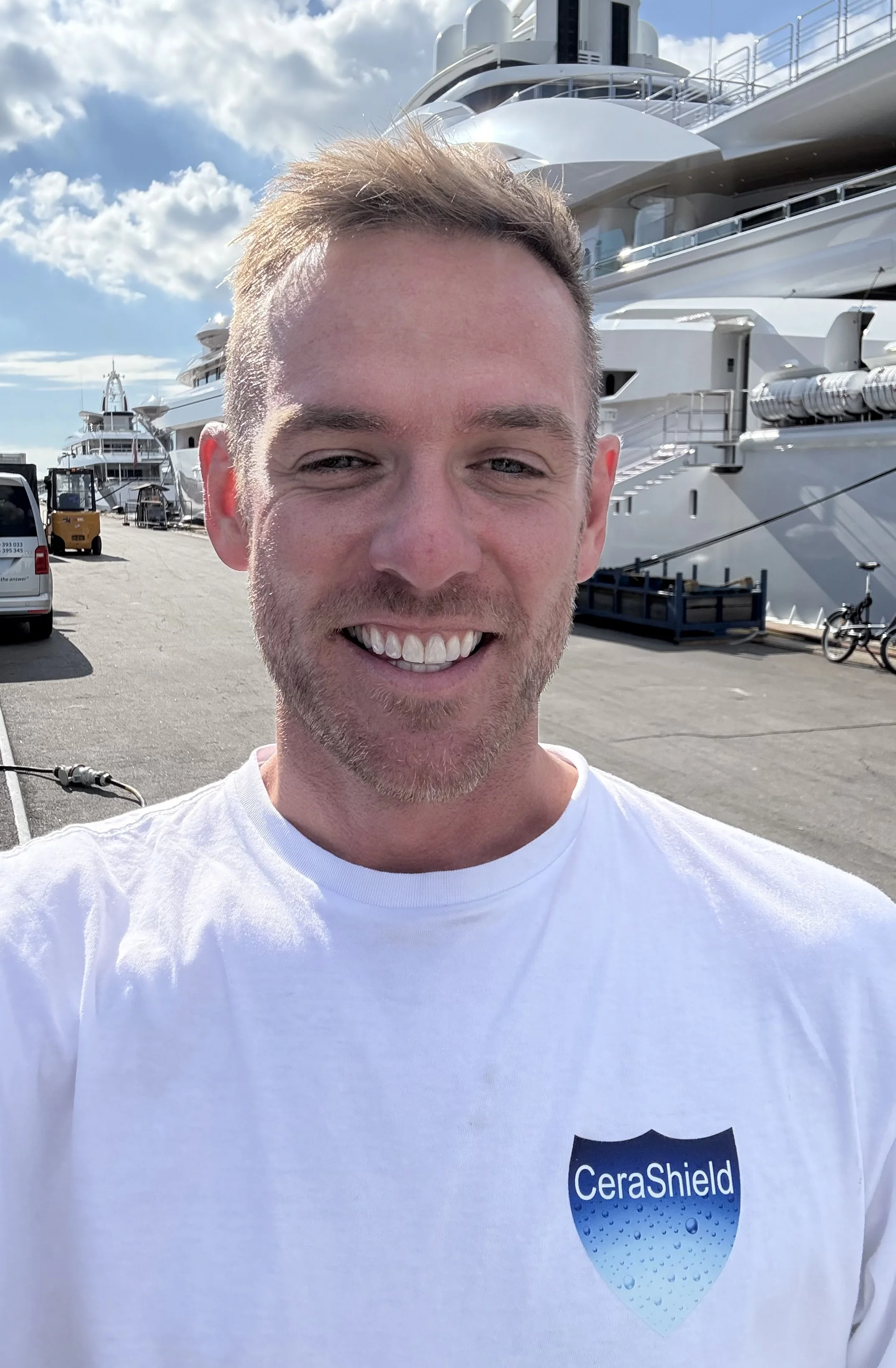 A man with short, light brown hair and a beard, wearing a dark jacket over a tan shirt, stands outdoors with boats and a marina in the background.
