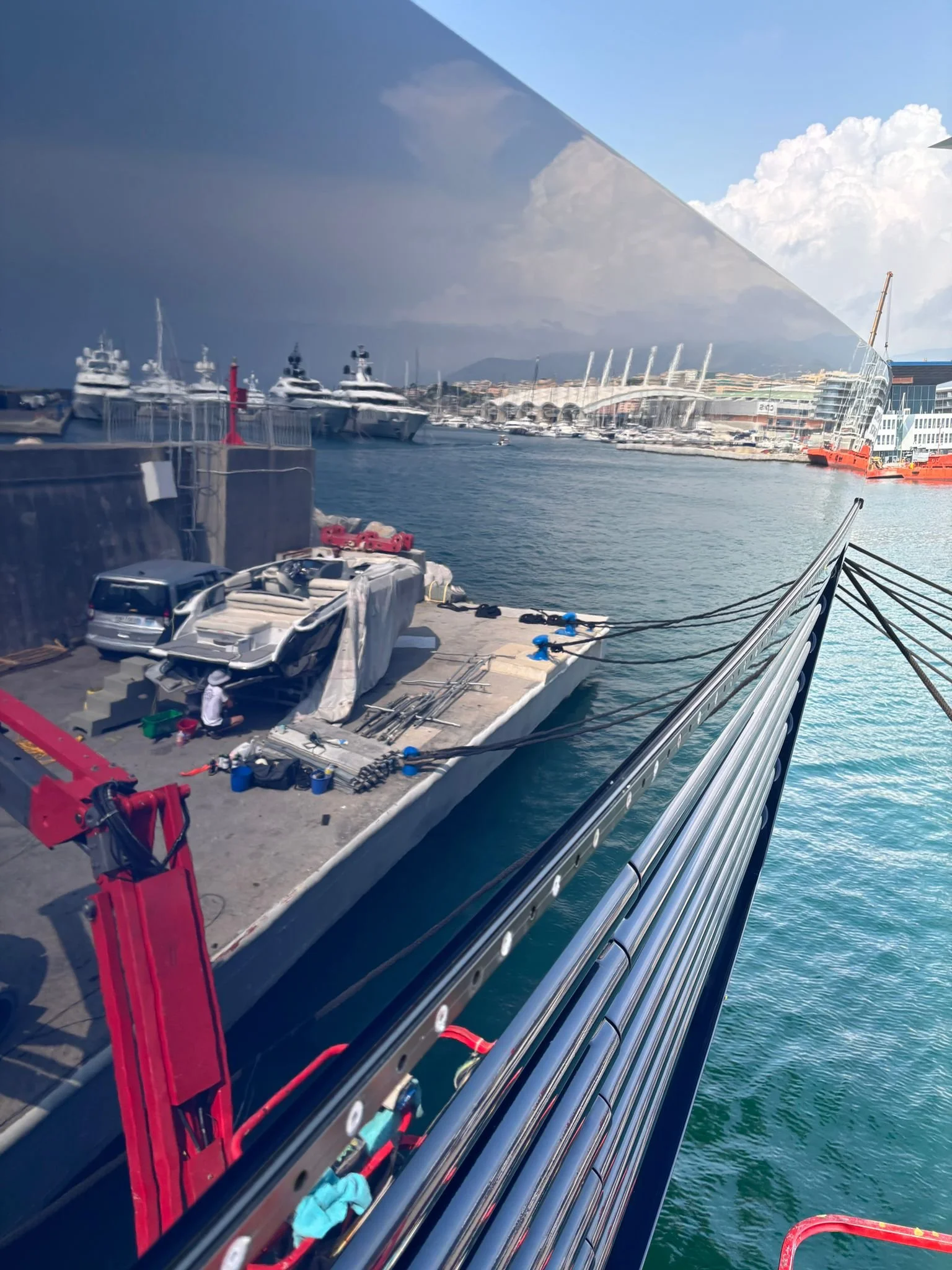 View of a harbor with yachts, a large luxury boat being serviced on the dock, and buildings in the background, with a reflective glass surface mirroring the scene.