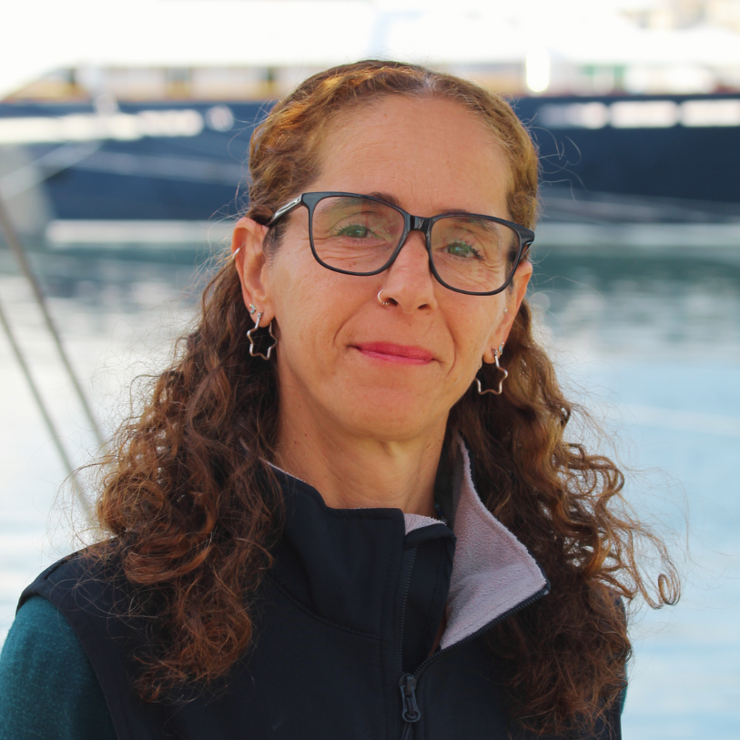 A woman with curly red hair, glasses, star-shaped earrings, and a nose ring, standing outdoors near boats on the water.