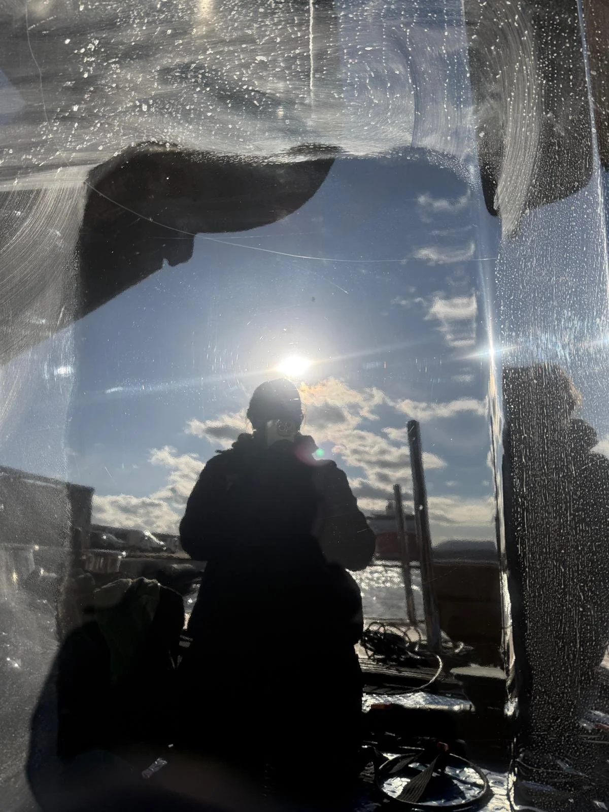 Reflection of a person taking a photo outdoors near water, with the sky, clouds, and the sun visible in the background, seen through a glass surface with water spots.