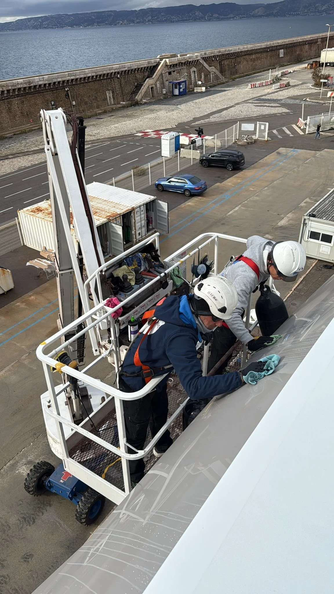 Two workers in safety gear are cleaning or inspecting the surface of an aircraft at an airport, using a scissor lift for access.