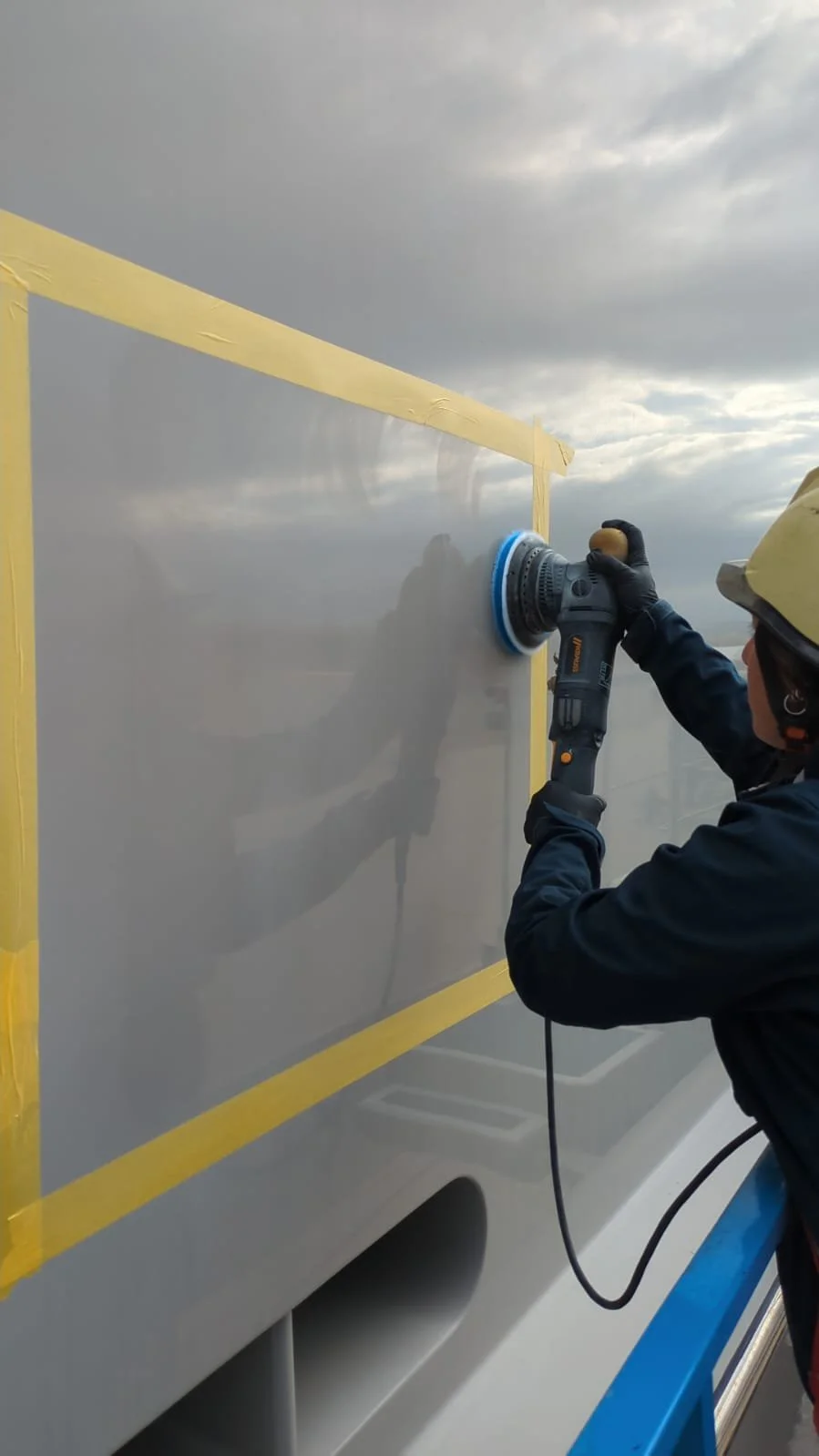 Worker in safety gear polishing a gray metal surface with a power buffer against cloudy sky background.