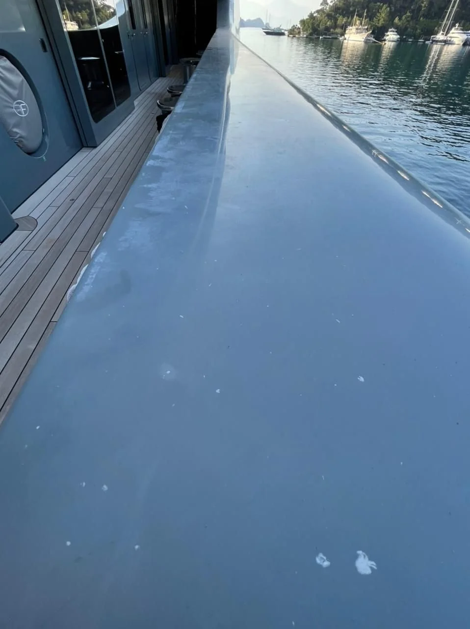 A yacht deck with a polished metal railing along the water's edge, and calm water with boats and trees in the background.
