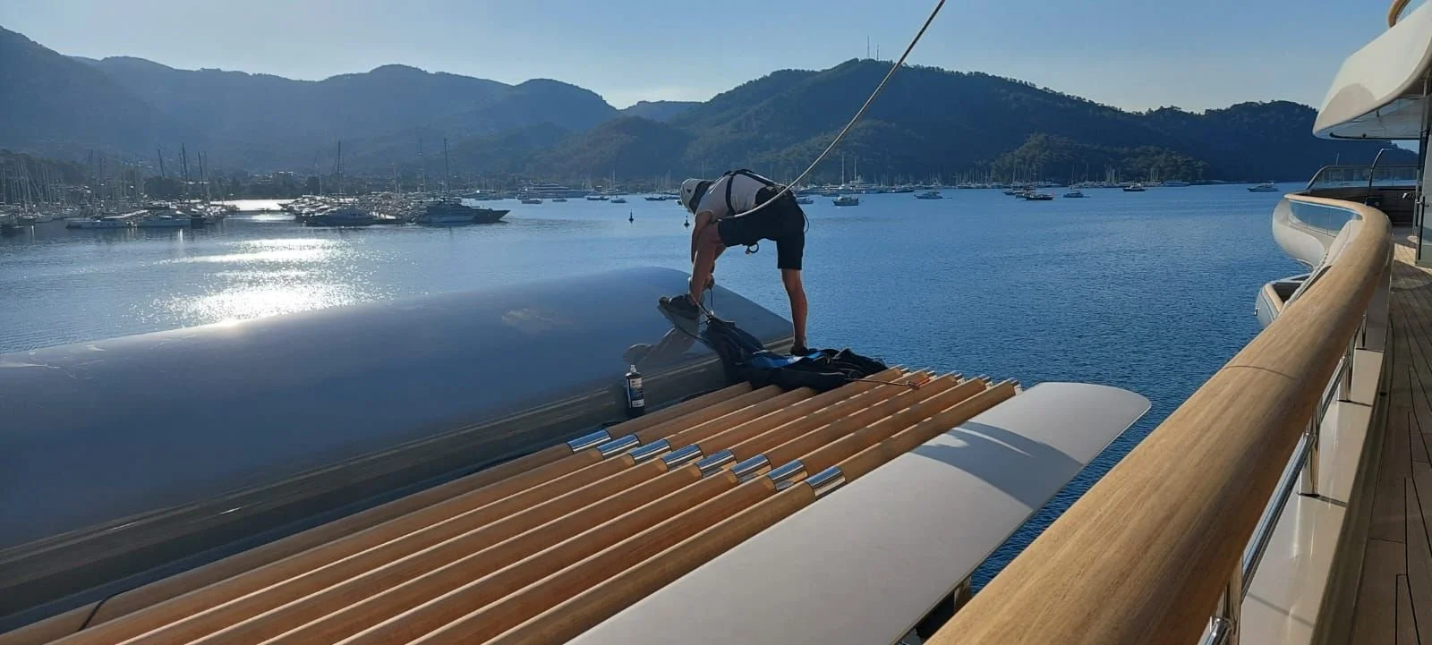 A person on a yacht deck securing a large inflatable boat to the yacht's surface against a marina with boats and mountains in the background.