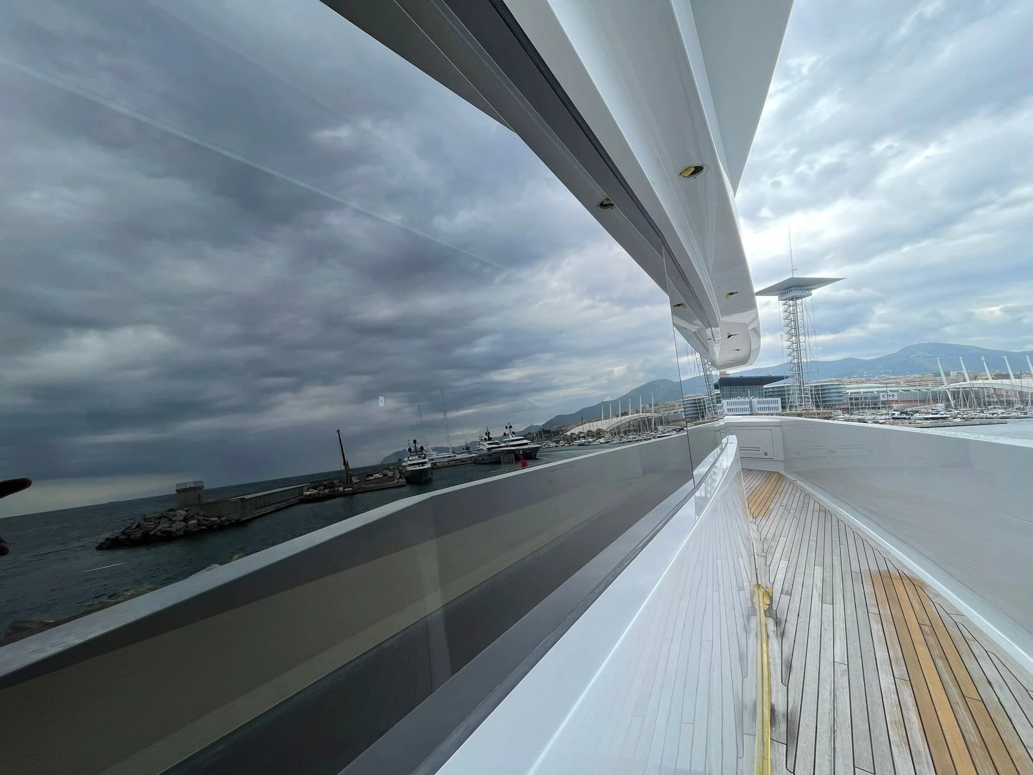 View from a yacht showing a marina with boats, overcast sky, and mountains in the background, with reflection of the yacht's exterior in a large window.