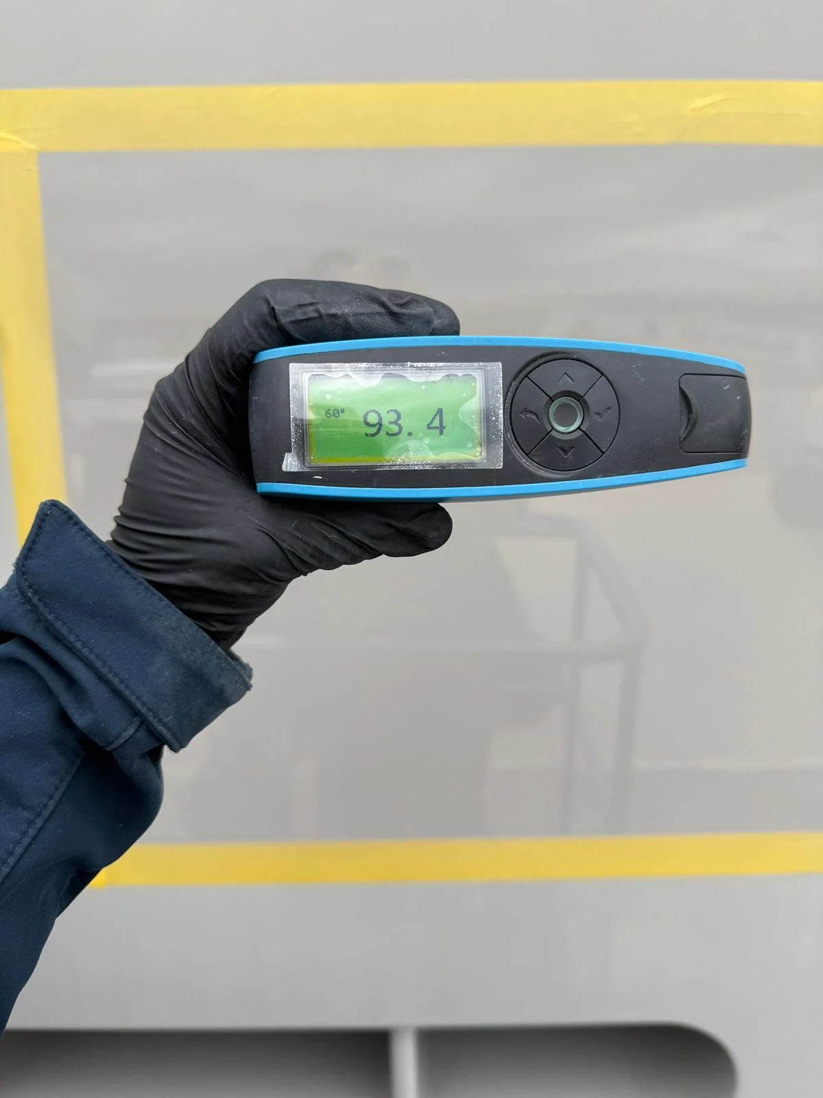 A person wearing a black glove holds a handheld device displaying a reading of 93.4. The background features a gray surface with a yellow caution line.
