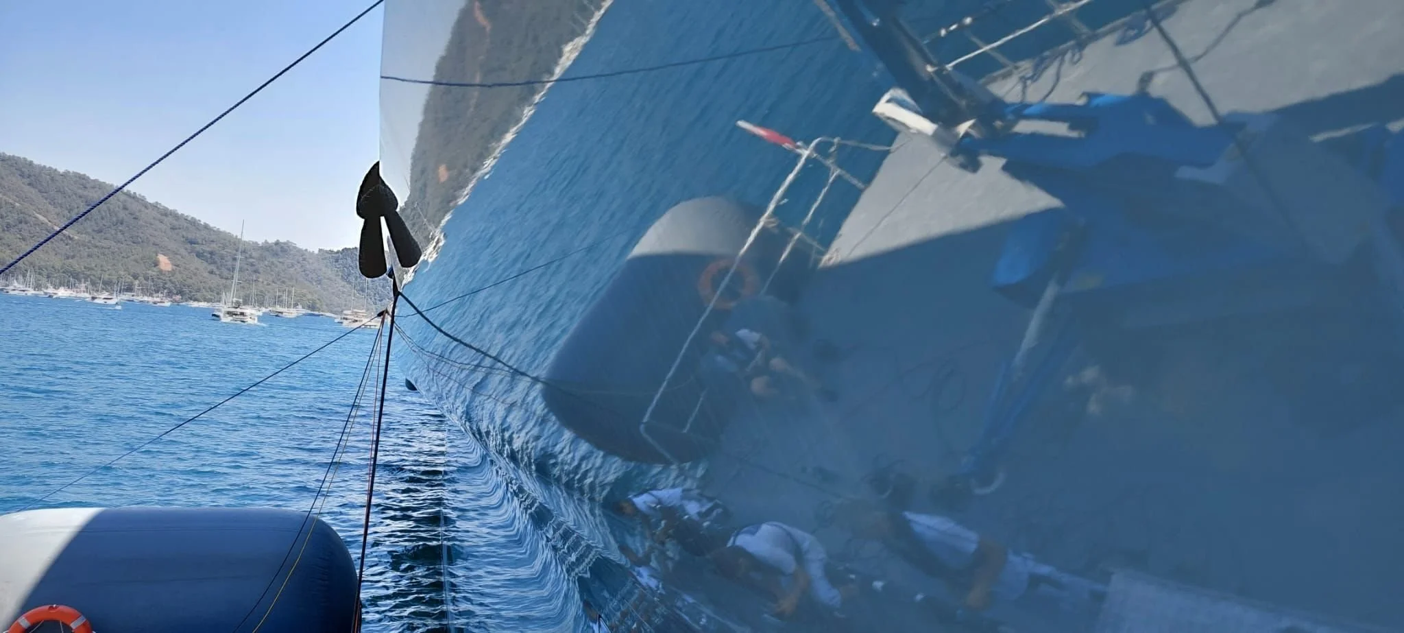 View of a sailboat from the side, showing the reflection of the boat and water with hills and other sailboats in the background.