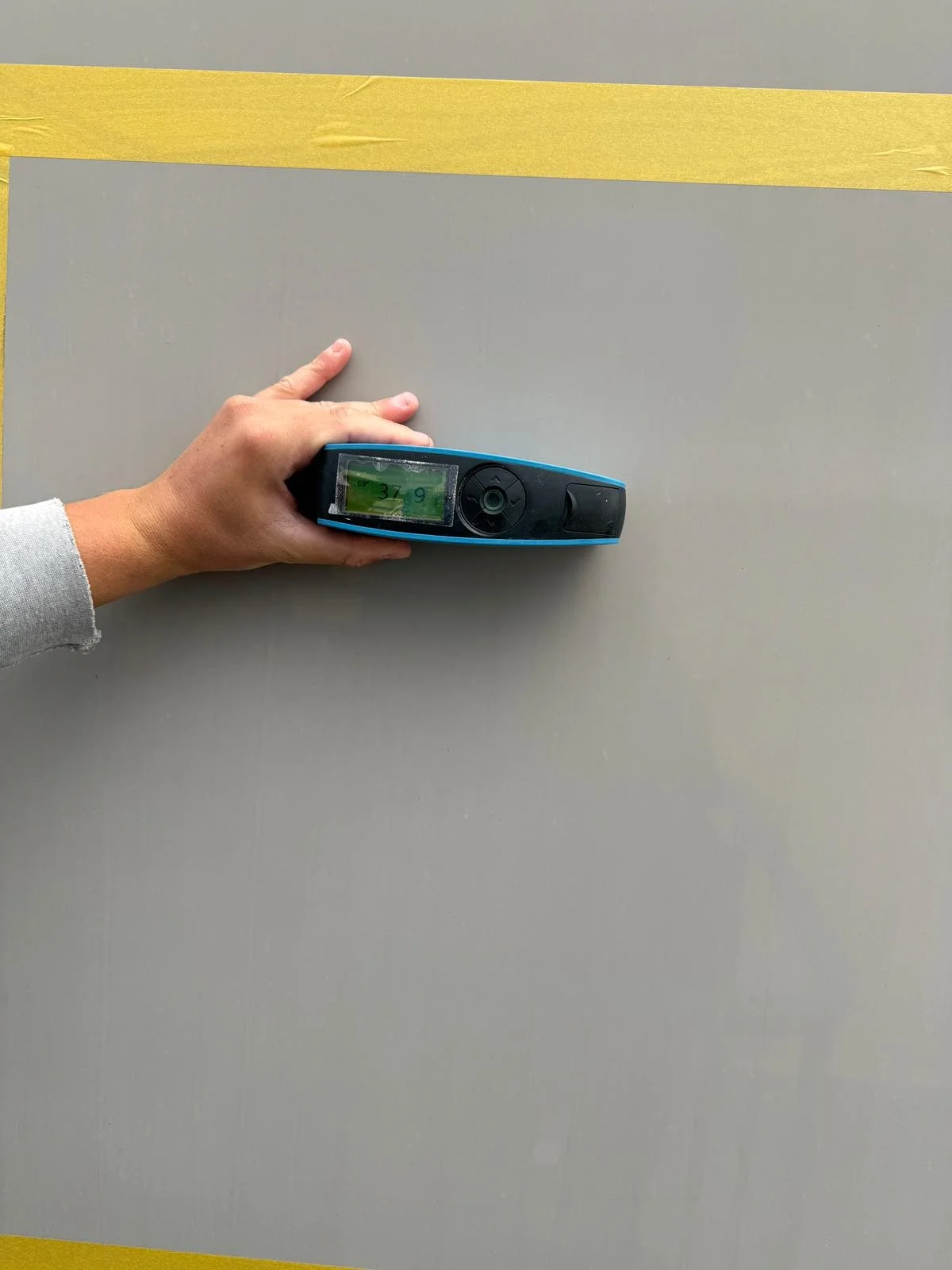Person holding a blue digital device with a green screen displaying the numbers 37.9 on a gray surface with yellow tape.