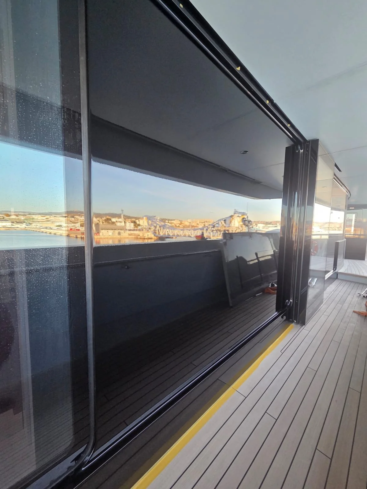 View from a balcony showing a large glass sliding door, outdoor deck with wood paneling, and a cityscape with buildings and a bridge in the distance during daylight.