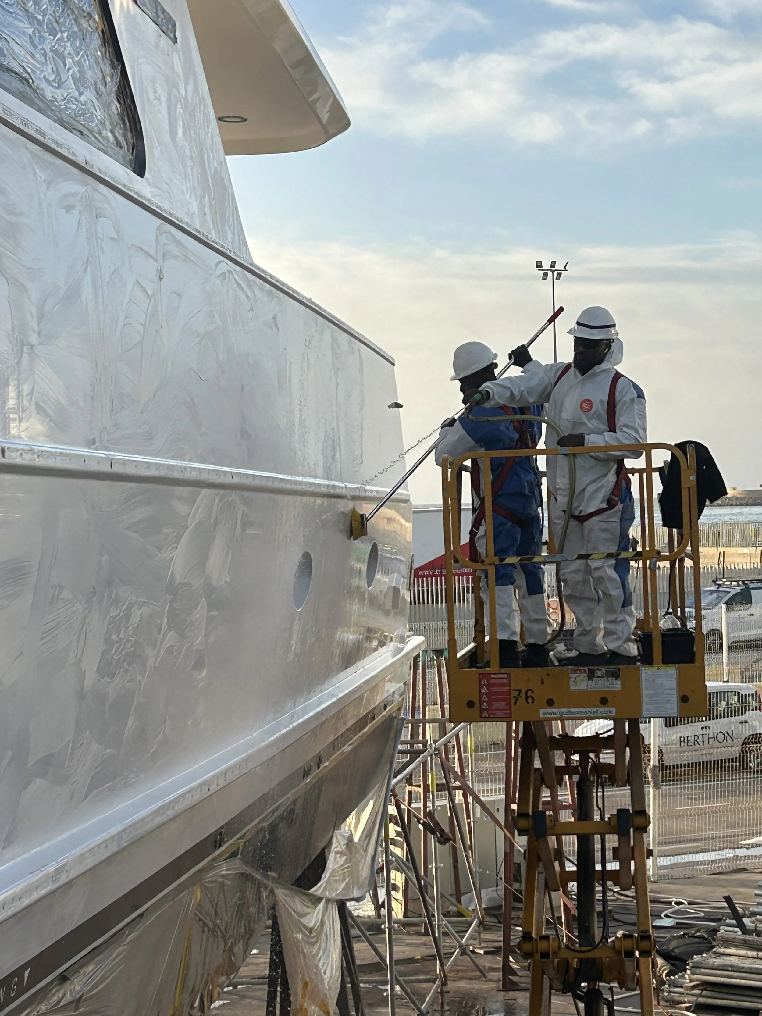 A worker wearing a white helmet and protective coveralls is sanding or polishing a blue surface inside an industrial setting.