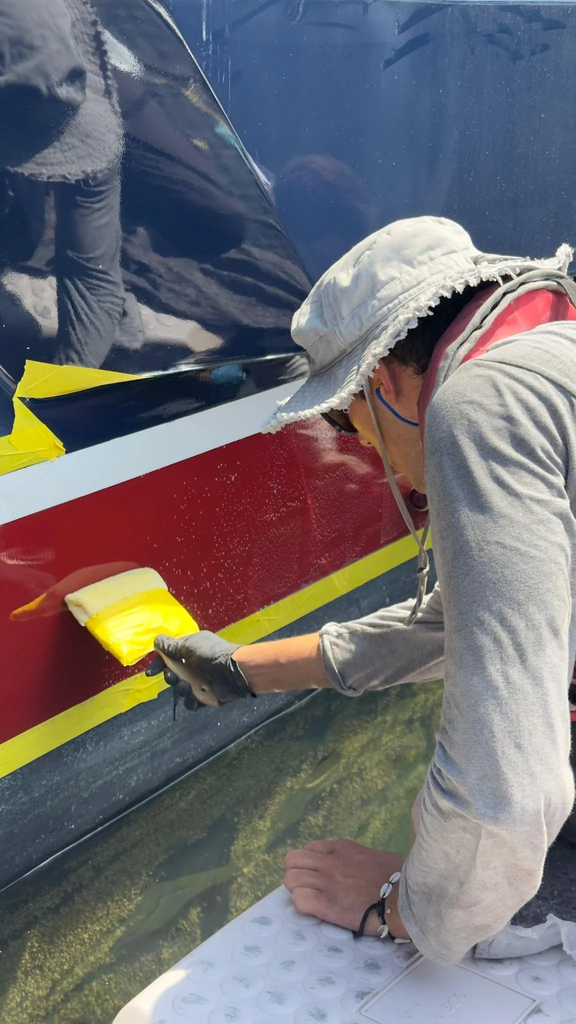 Person painting a boat with a yellow brush, wearing a sun hat, sunglasses, and gloves.
