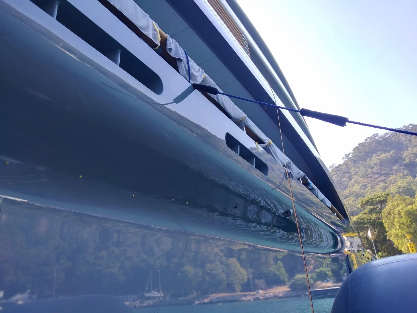Close-up view of a yacht's hull reflecting the surrounding trees and sky, with part of a mountain in the background, and a ladder or rope attached to the side.