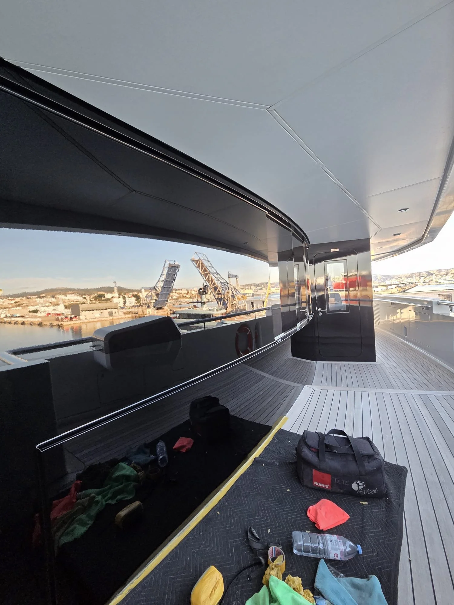 The image shows a modern rooftop outdoor space in a city, with a black, shiny railing and a smooth, light-colored flooring. Scattered on the floor are various items, including a bottle of Evian water, colorful clothing or towels, backpacks, and a yel