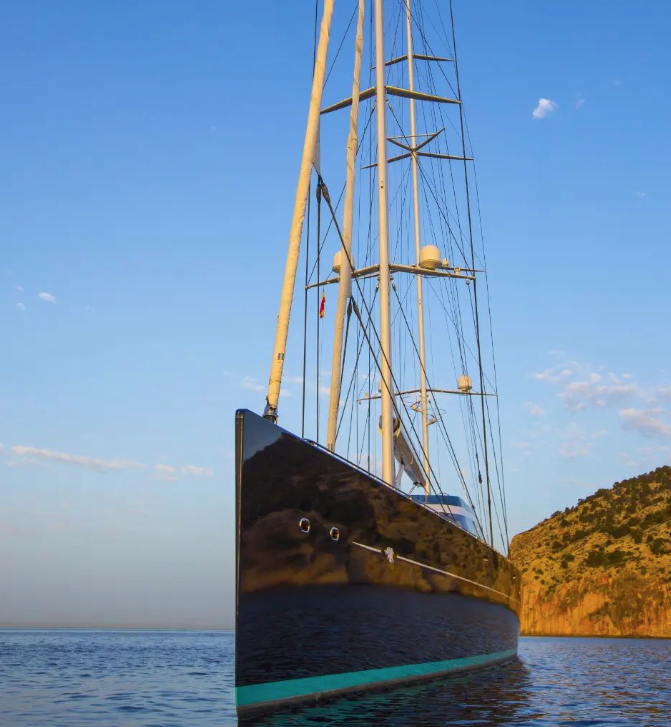 A large sailing yacht with tall masts and sails docked on calm water near a hillside at sunset.