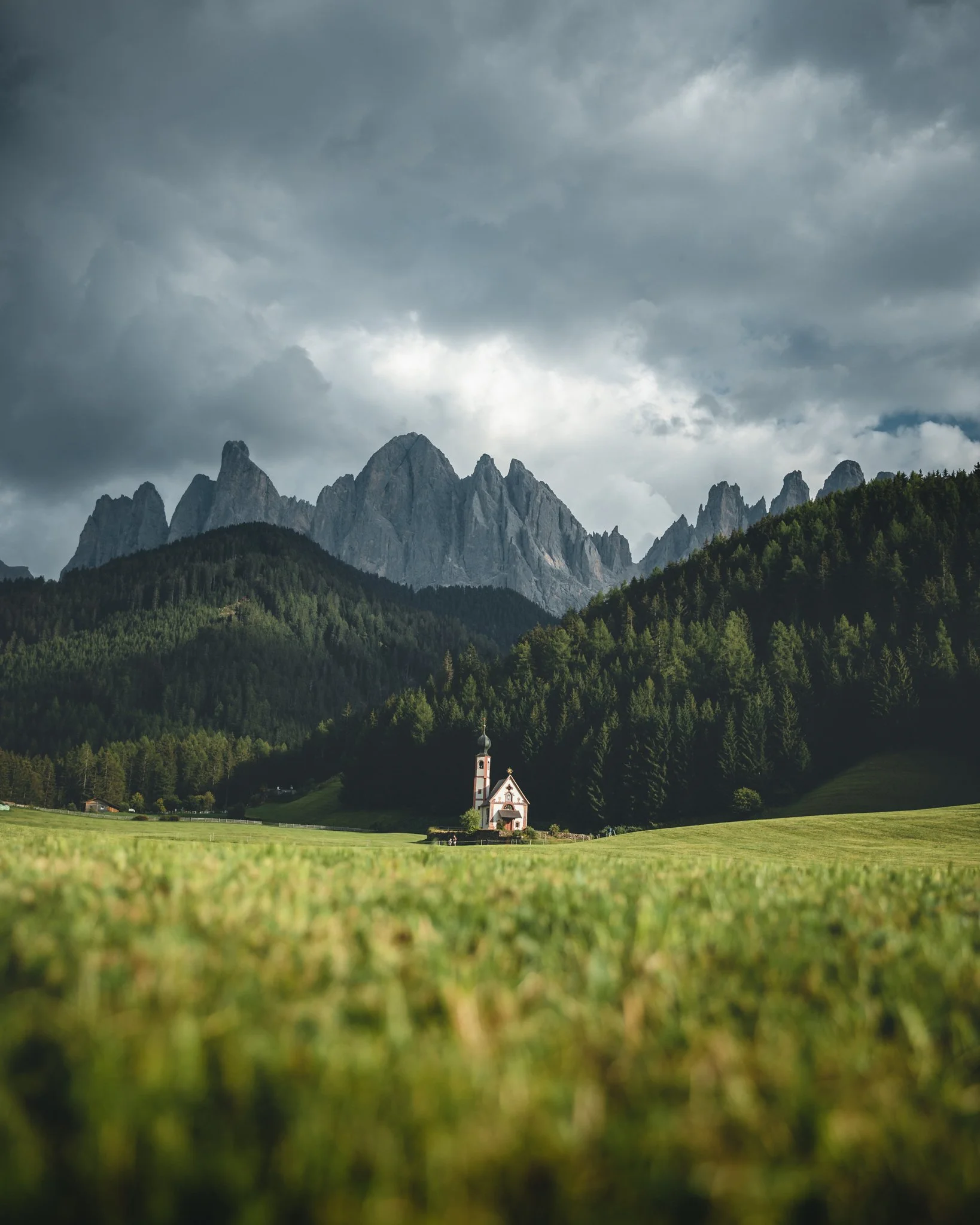 A small church with a steeple is situated in a grassy field, surrounded by dense green forests, with rugged mountain peaks and dark clouds in the background.