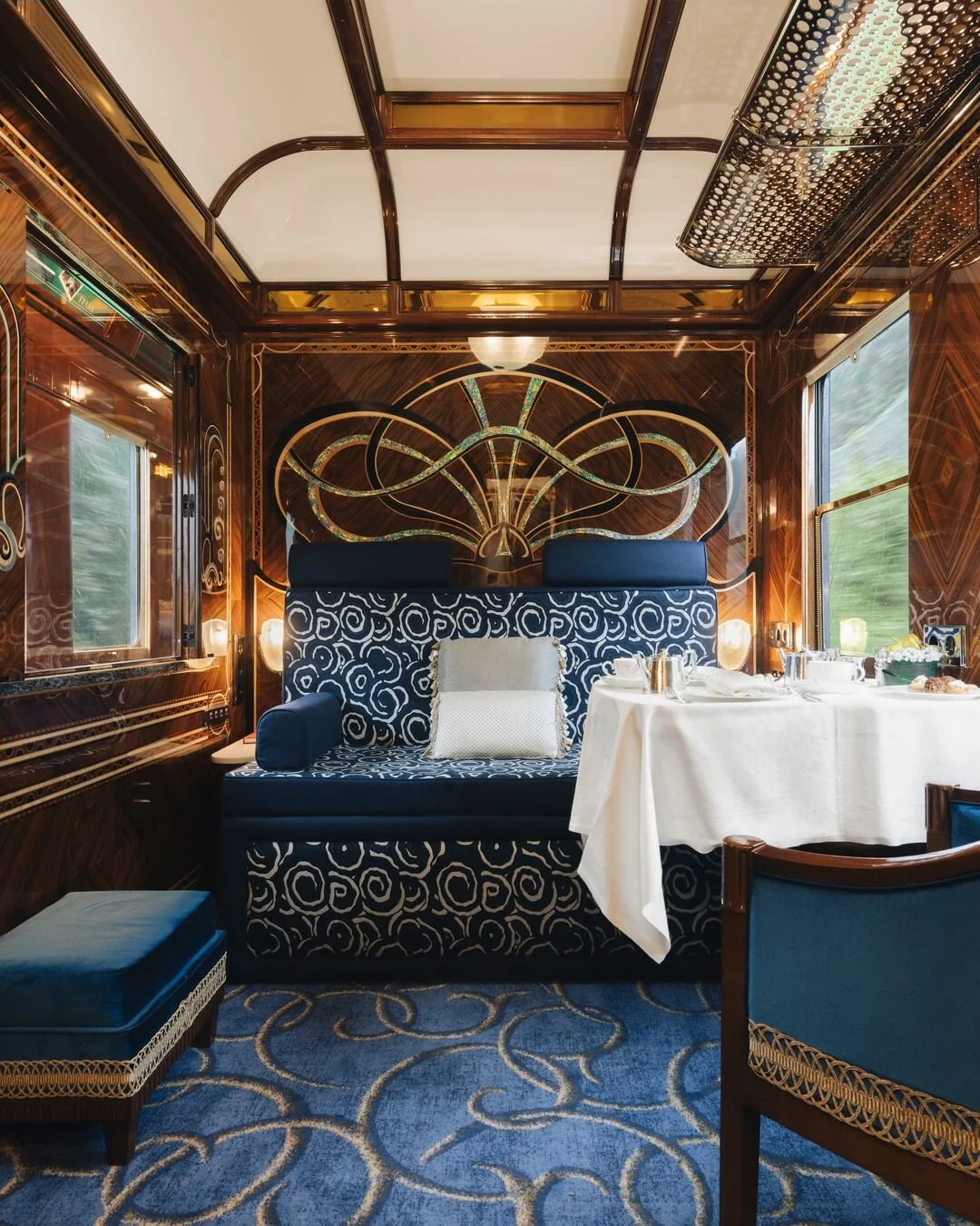 Luxurious dining area on the Venice Simplon-Orient-Express with dark wood panelling accented with gold and glass designs, a plush patterned blue and white sofa with a pillow, a table set with white tablecloth, and silverware.