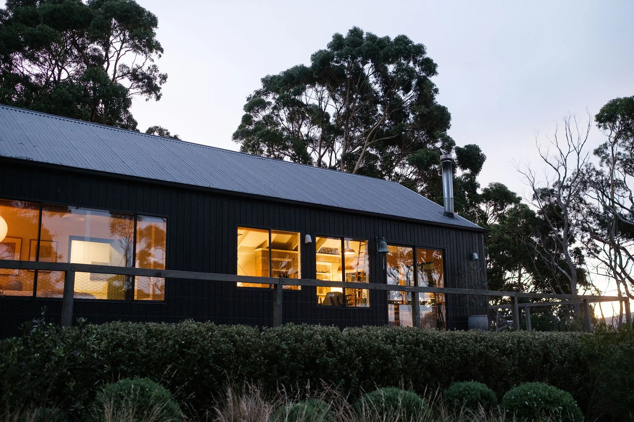 A house with black walls and large windows with warm interior lighting, situated on a hillside surrounded by trees and bushes, during dusk.