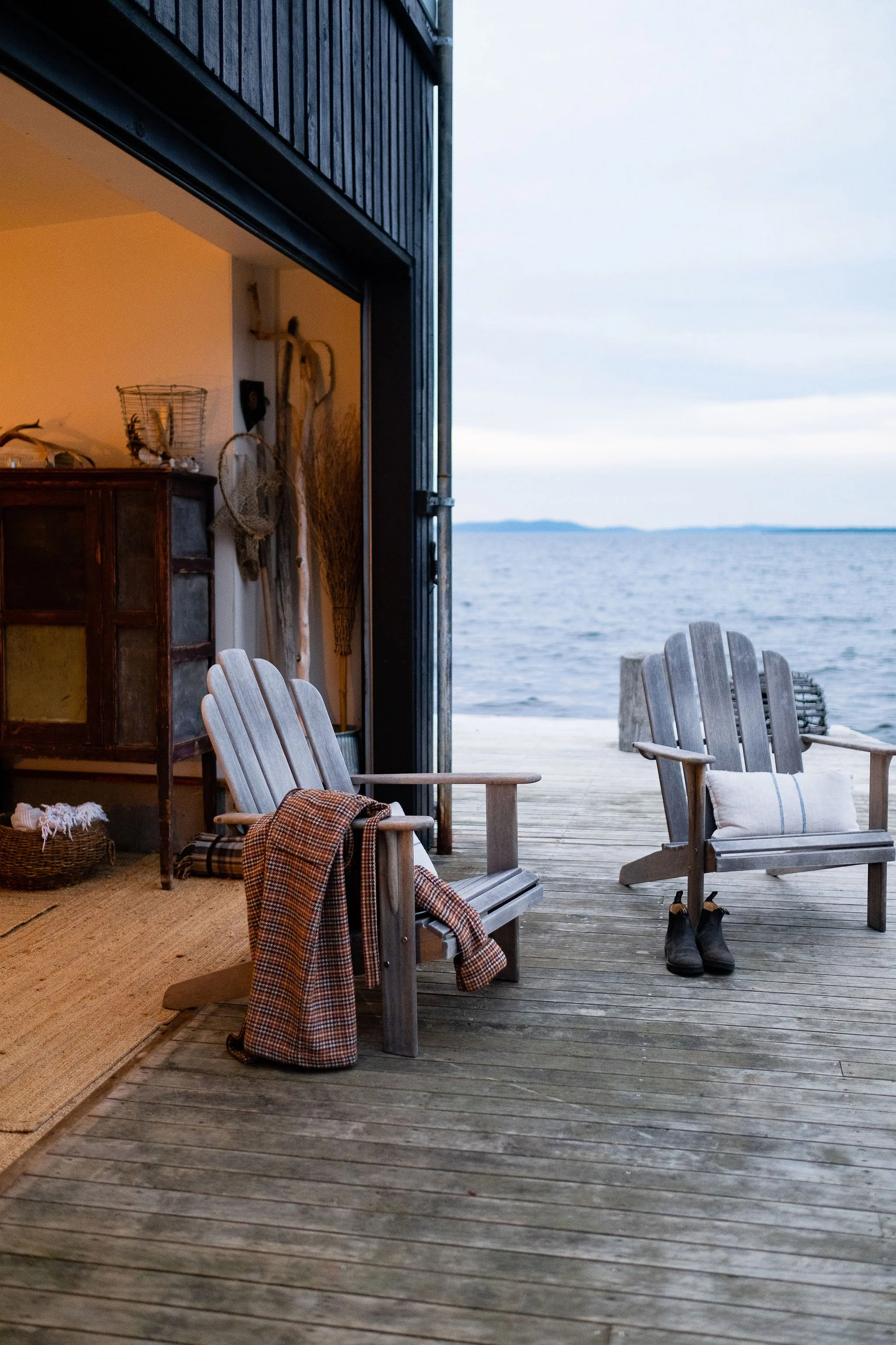A cozy outdoor wooden deck with two Adirondack chairs overlooking a body of water, with shoes placed underneath one chair and a blanket draped over the other, while the interior of a home with warm lighting is visible through a partially open sliding door.