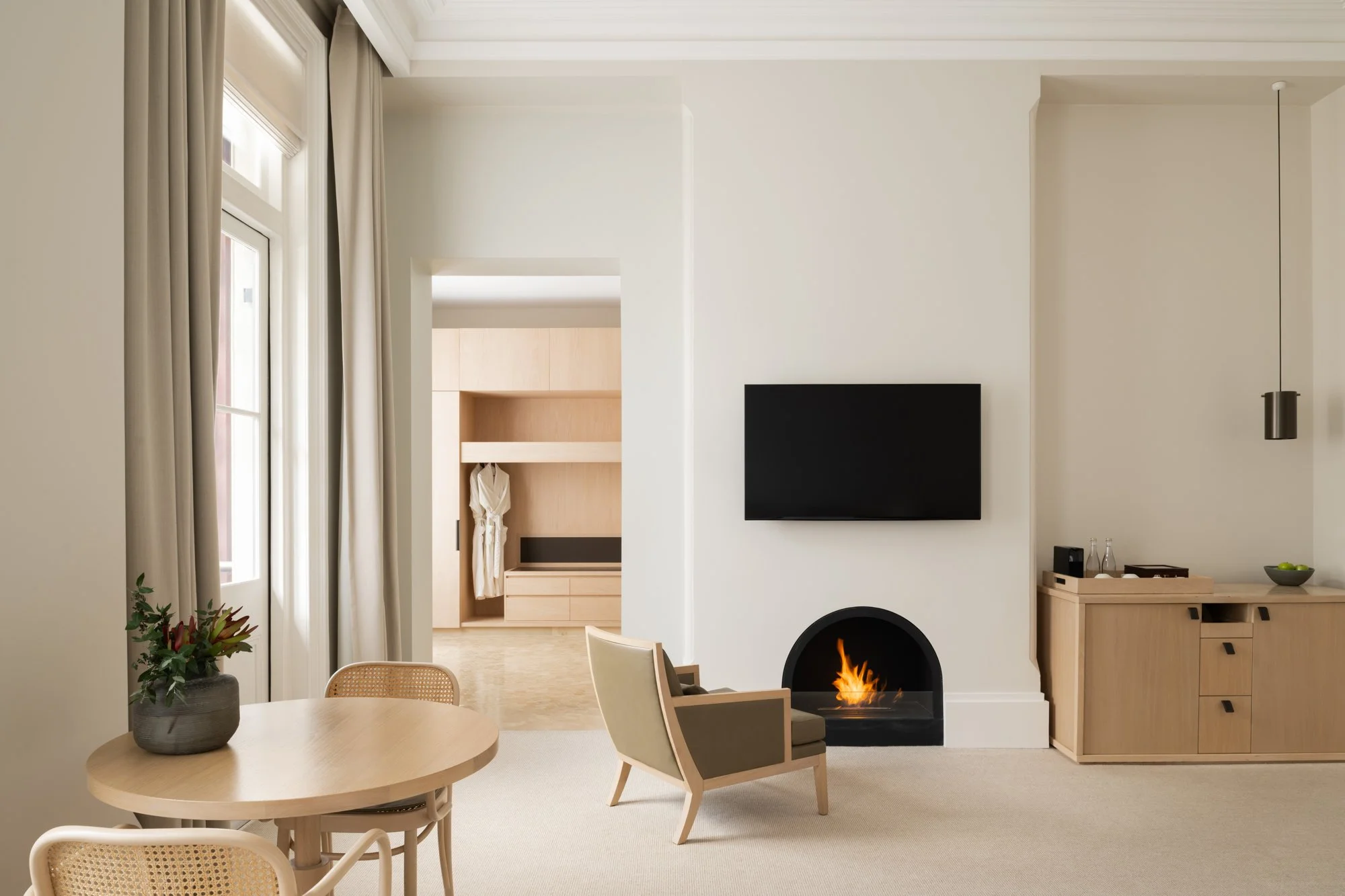 Living room with a beige theme, featuring a wooden round table with a flower arrangement, a chair with beige and brown upholstery, a wall-mounted flat-screen TV, a stove with a visible flame, a built-in cabinet with glass bottles and a bowl of green apples, and large windows with beige curtains.