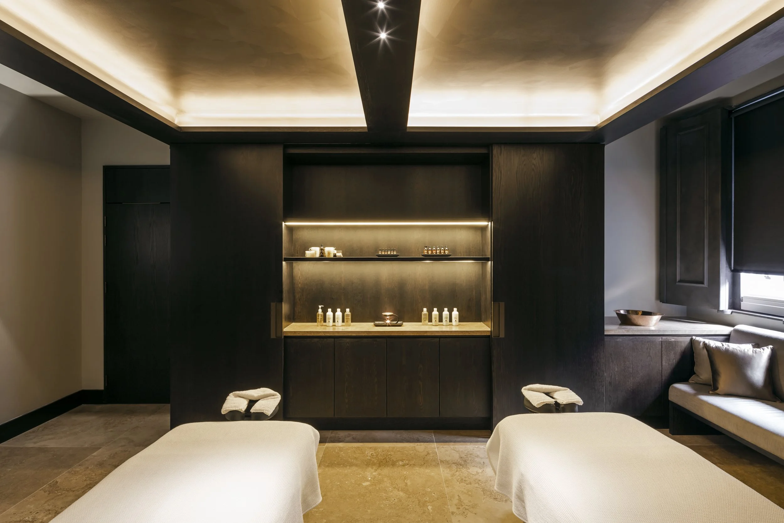 Spa room with two massage tables, dark wood cabinet with bottles and skincare products, and a window with black shade