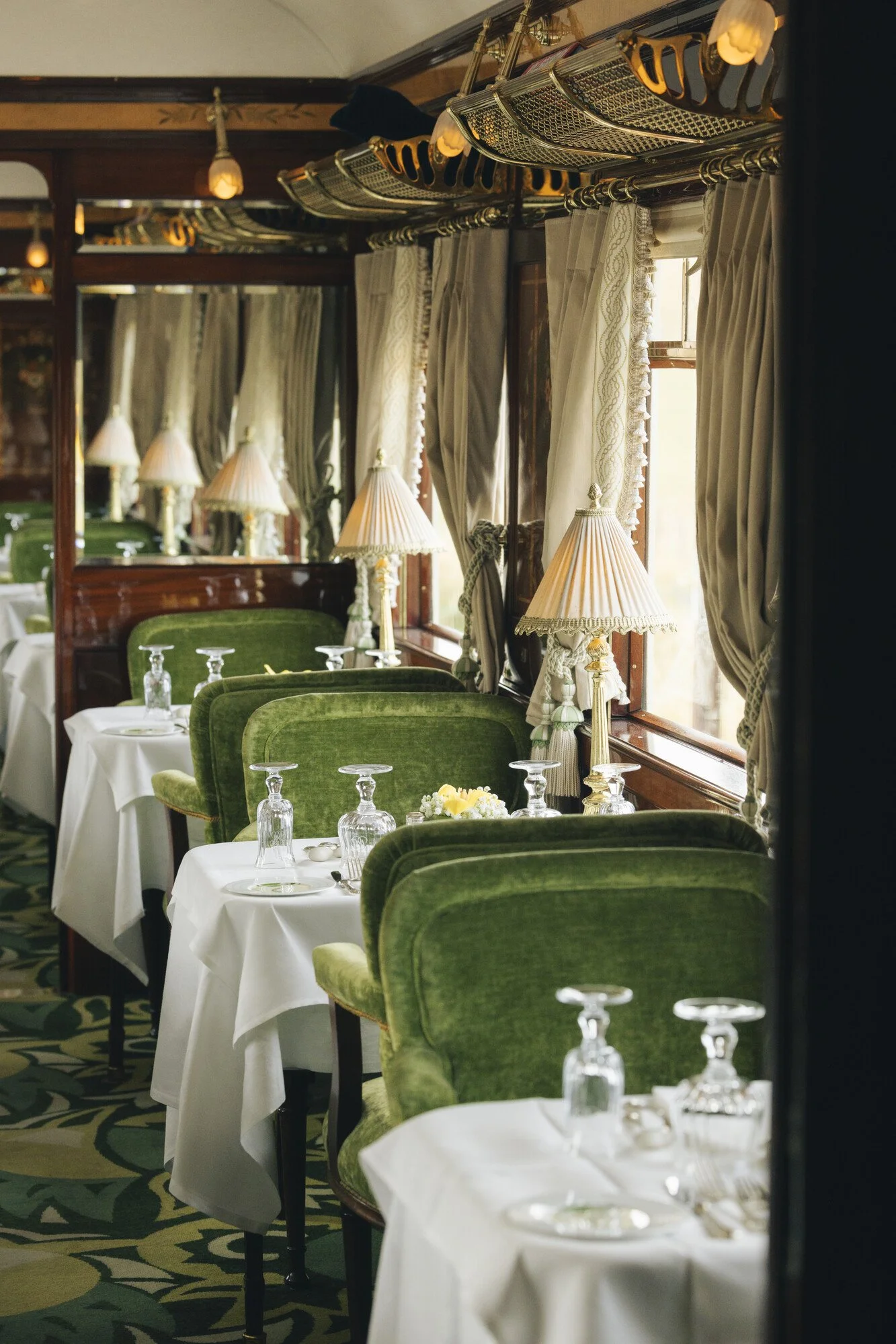 Elegant dining room with white tablecloth-covered tables set with upside-down glasses and small floral centerpieces. Green plush chairs surround the tables. Tall windows with lace curtains allow natural light, and classic table lamps with pleated sha