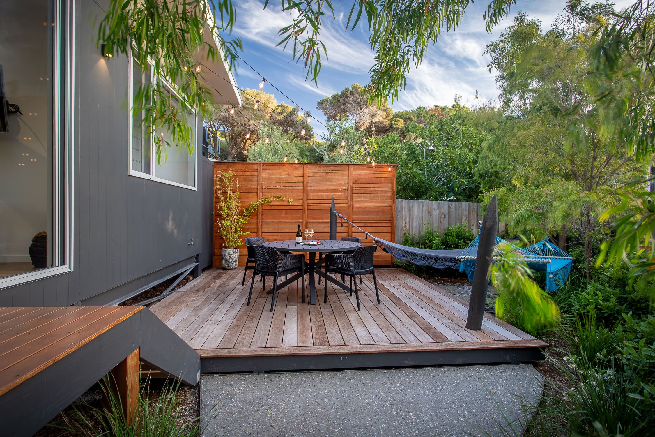 Outdoor patio with wooden deck, black table, four black chairs, two wine bottles, glasses, hammock, potted plant, string lights, surrounded by trees and greenery.