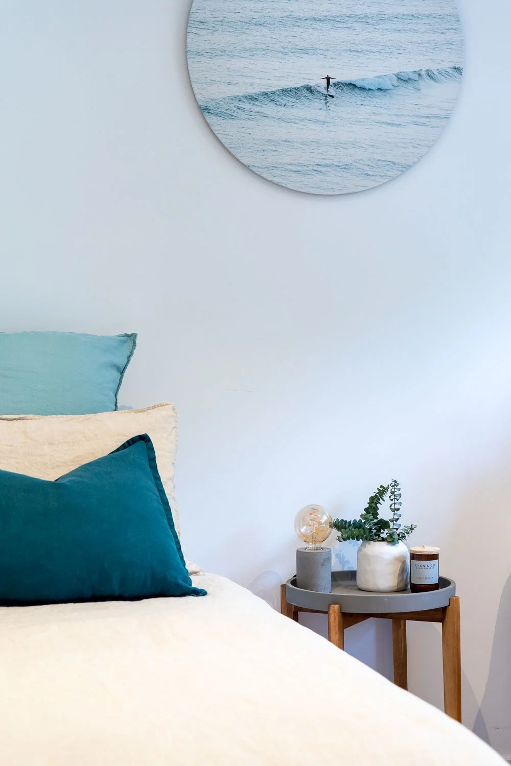 A bedroom with a white bed and teal pillows, a small wooden side table with a potted plant, a candle, and a decorative light bulb, and a round mirror on the white wall reflecting a person surfing on the ocean.