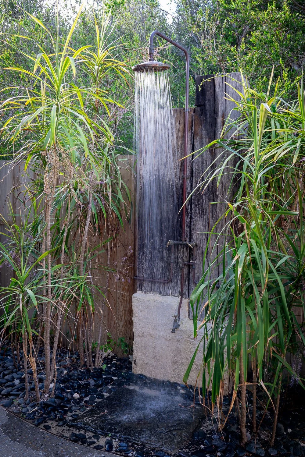 Outdoor shower with running water, surrounded by lush green plants and black stones on the ground.