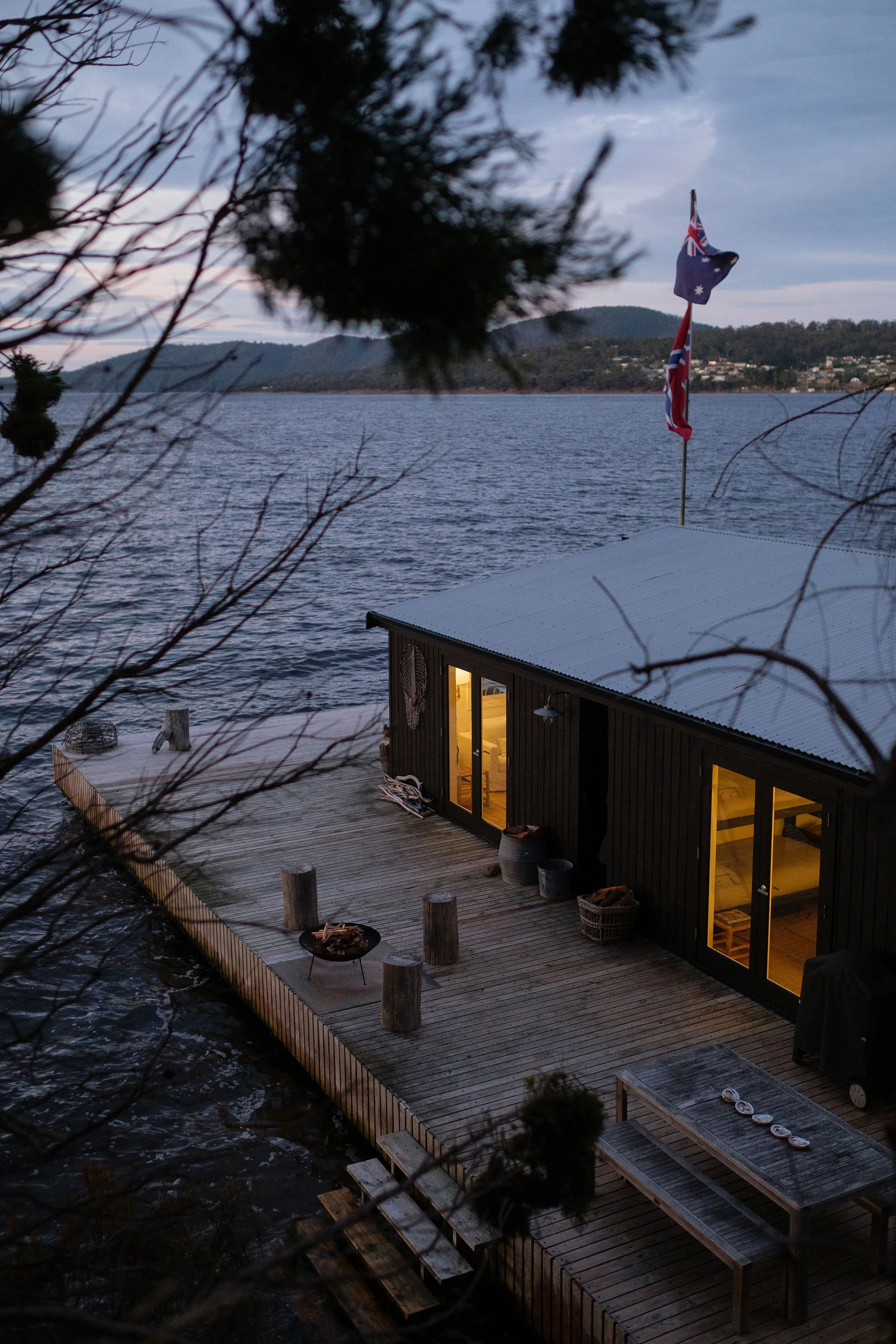 A lakeside house on a wooden dock illuminated from inside, with Australian flags on the roof, surrounded by trees, and a calm body of water with distant hills in the background during dusk.