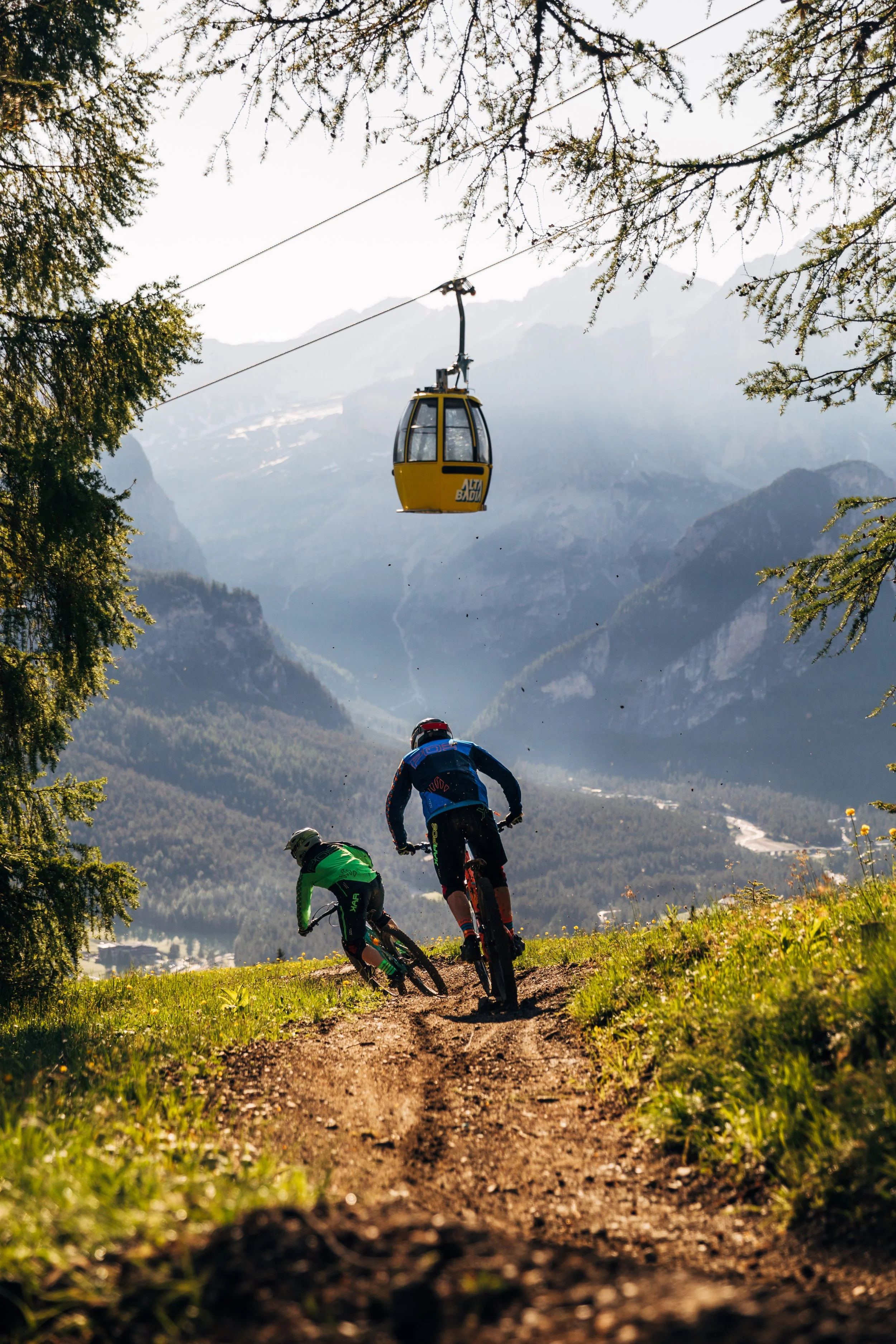 Two mountain bikers riding on a dirt trail in a mountainous area with a valley in the background, and a cable car suspended above them.