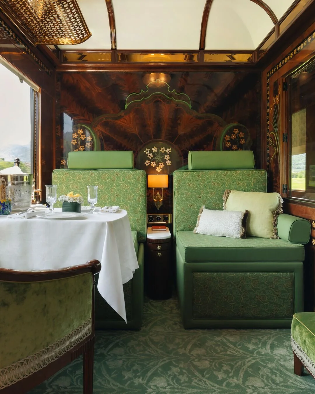 Elegant train dining car interior with green upholstered booths, a round table set with glassware and white tablecloth, and decorative wood paneling with floral motifs.