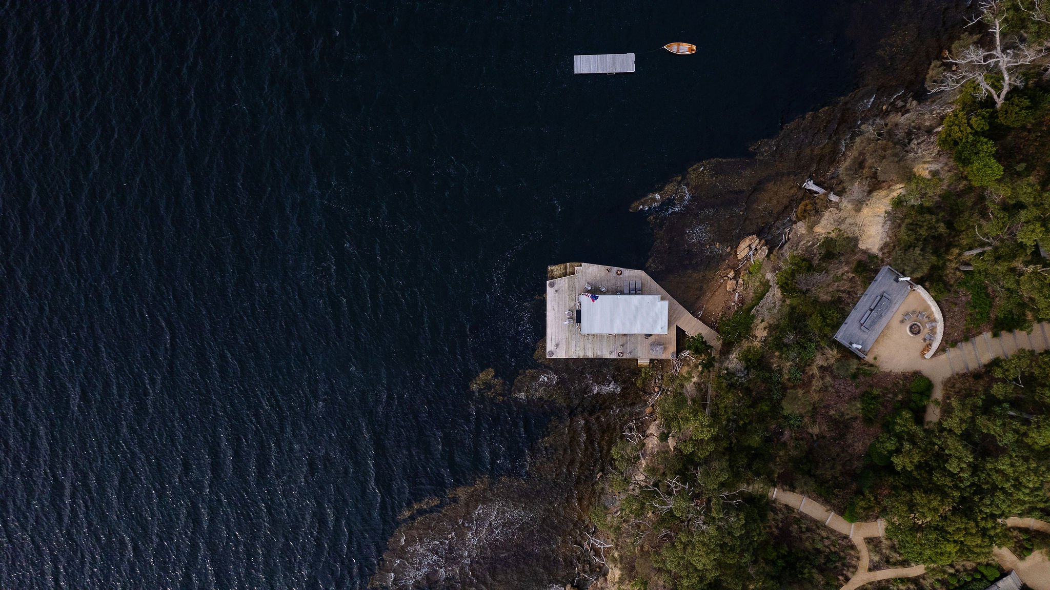 An aerial view of a coastal area showing a house on a cliff, a dock extending into the water, and a boat nearby.