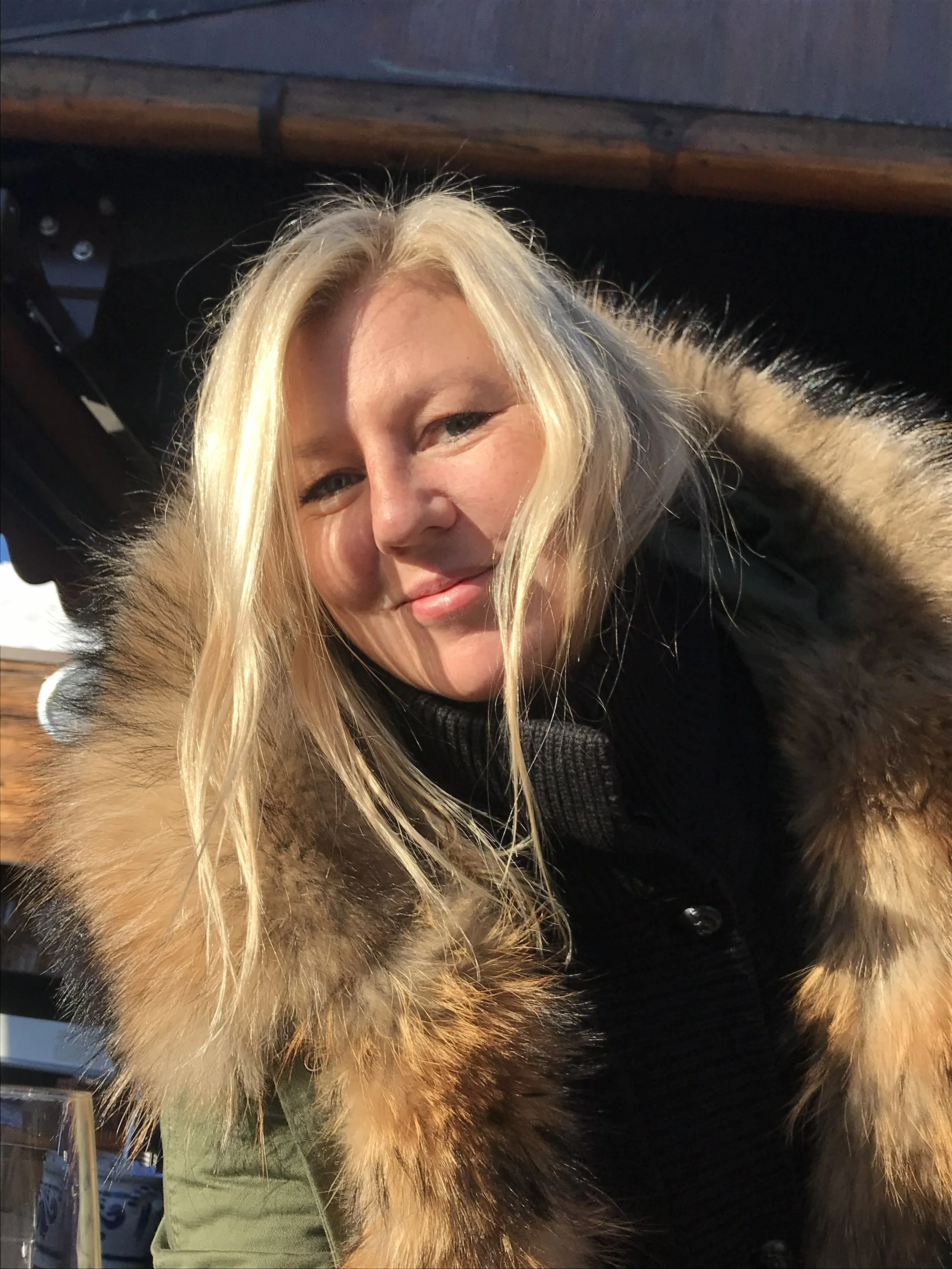 A woman with long blonde hair smiling outdoors, wearing a black sweater and a coat with a fur-lined hood.