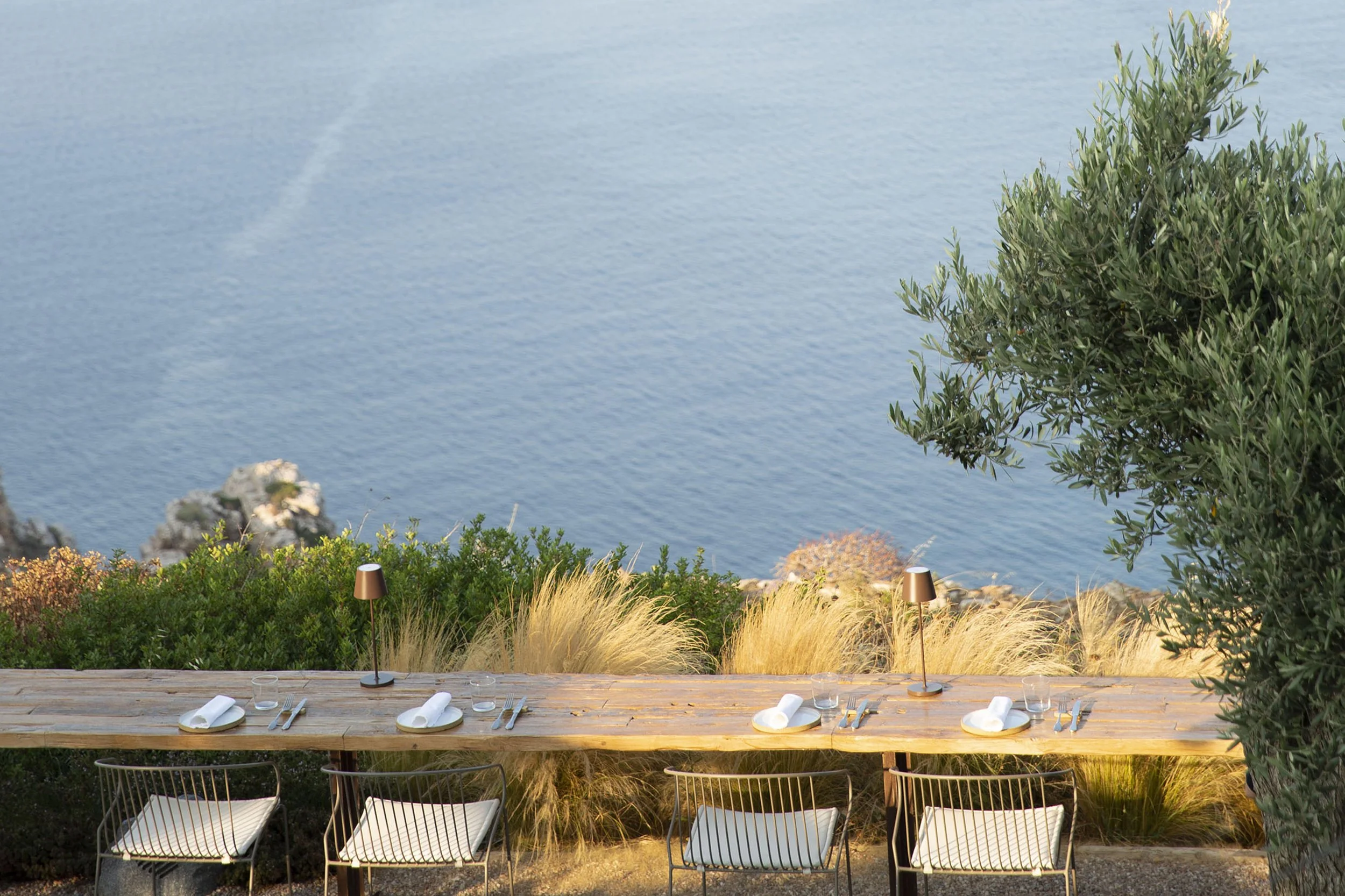 Outdoor dining table set with plates, glasses, cutlery, and small lamps, overlooking a body of water with rocks and bushes.