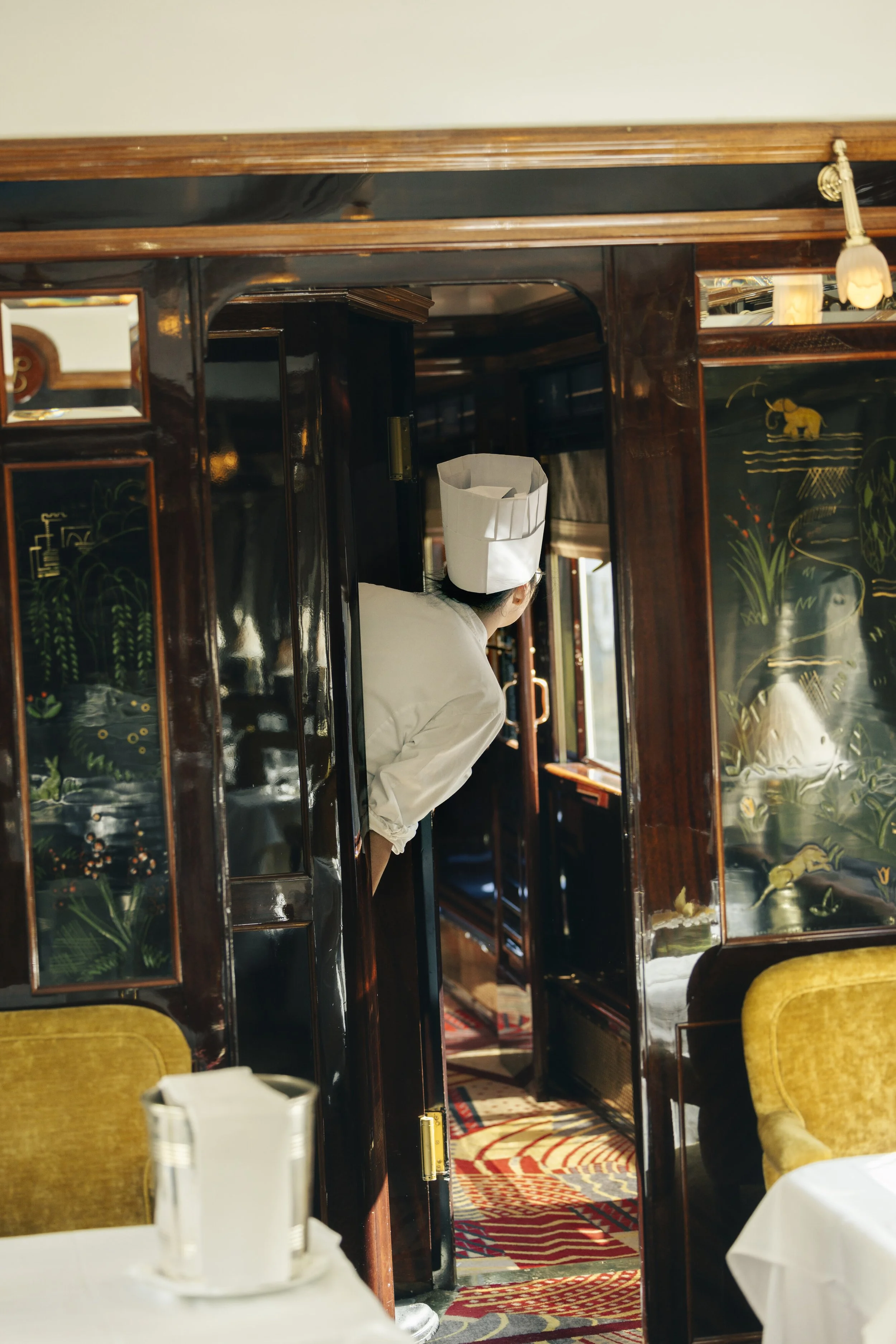 A chef peeking around a corner in a restaurant with dark wood paneling and decorative Asian-style paintings.