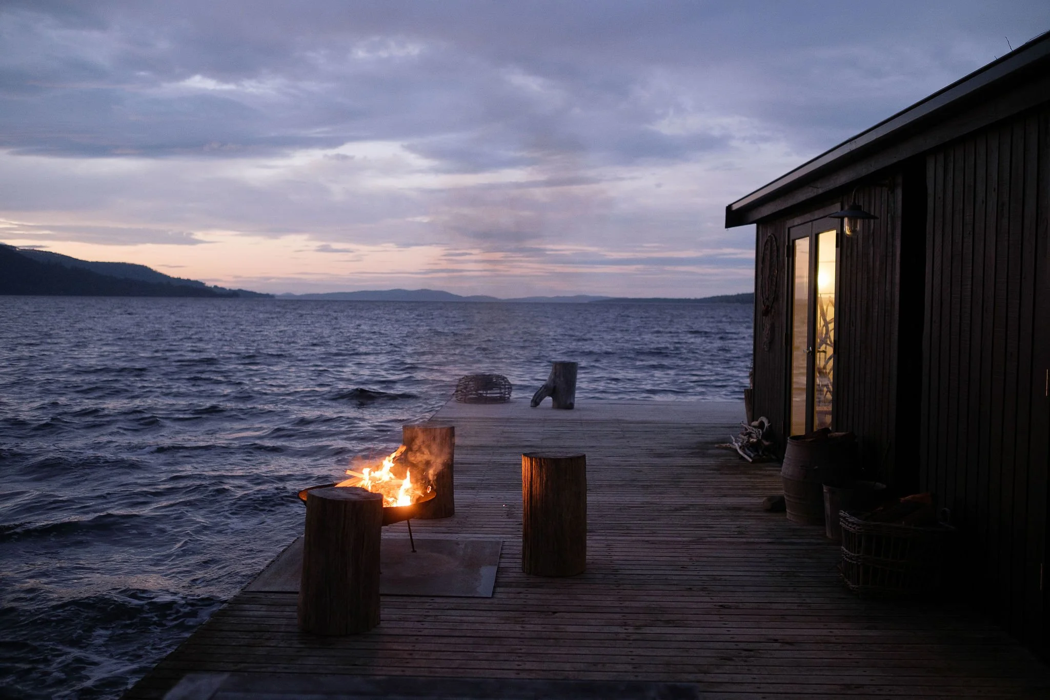 A wooden dock extending over a body of water during dusk, with a small fire burning between two tree stumps and a dark wooden house on the right side, reflecting the sunset in its window.