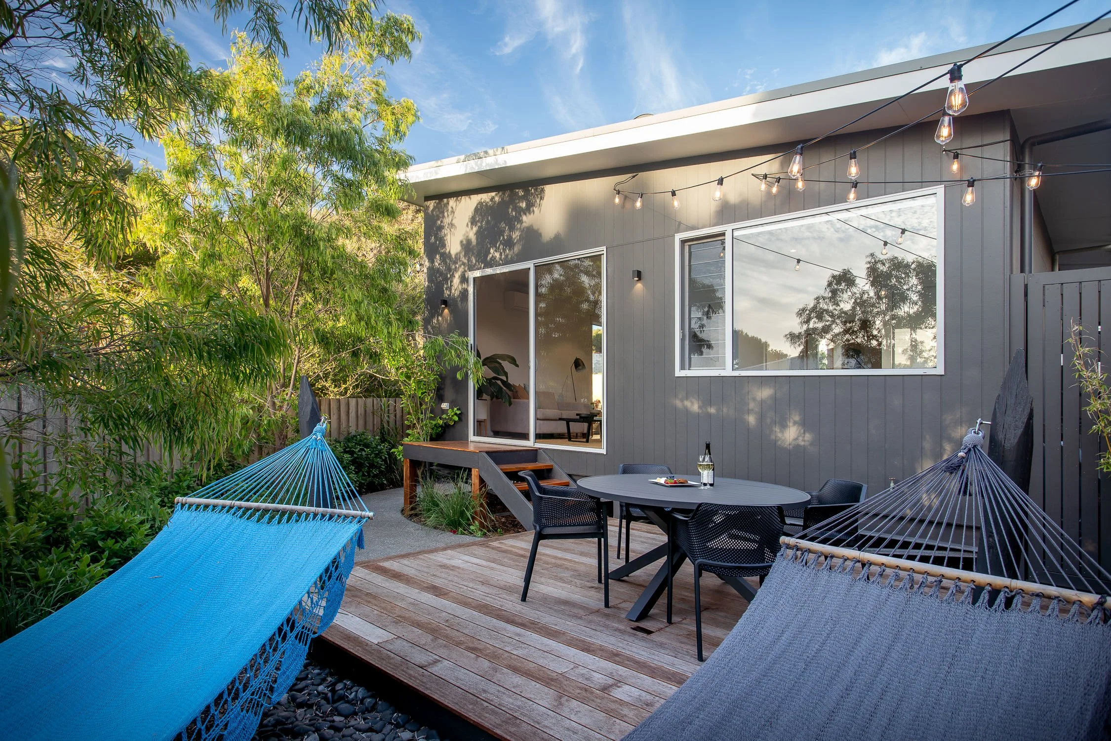 Outdoor patio with two blue hammocks, a black table with four chairs, string lights, and greenery in the background.