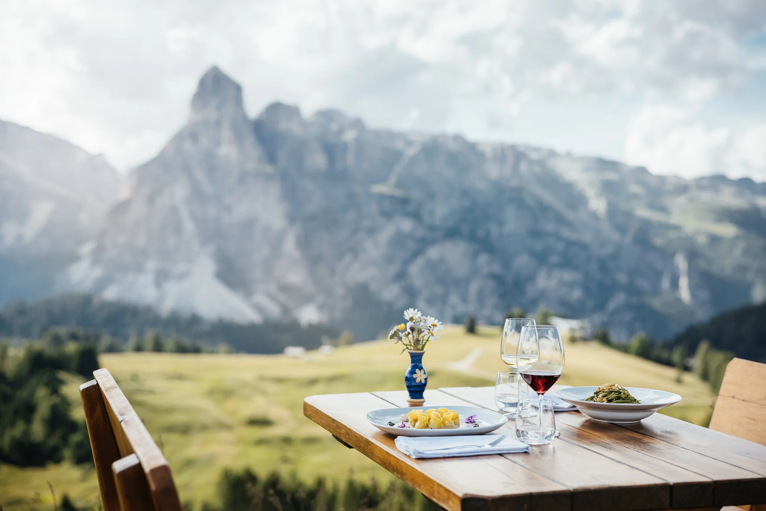 An outdoor dining table with plates of food, wine glasses, and a flower vase with daisies, set against a mountainous landscape with green fields.