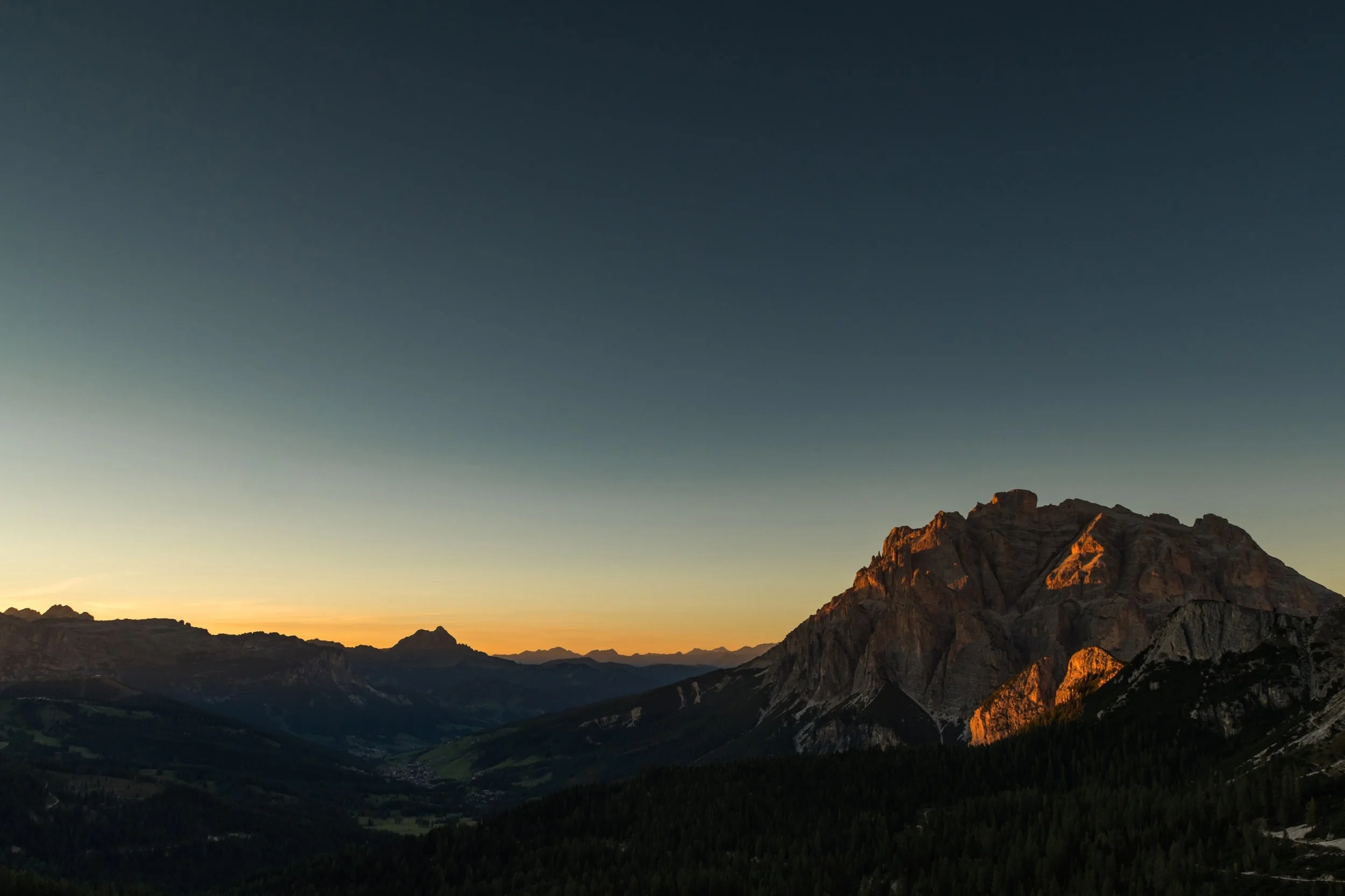 Sunset over mountain range with rocky peaks and forested slopes in the foreground.