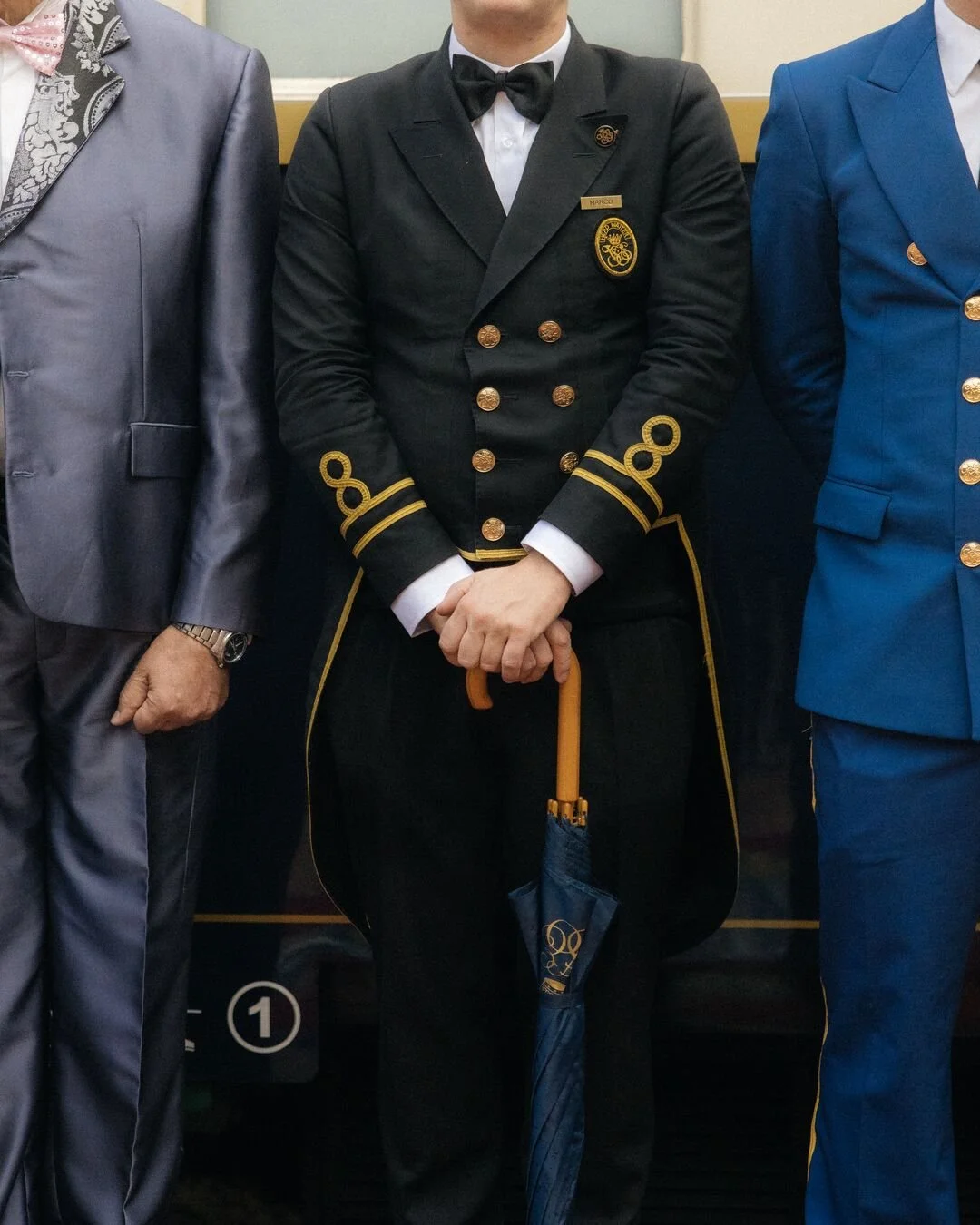 A person in a black formal uniform with gold accents and badges, holding a blue umbrella, flanked by two others in formal suits.