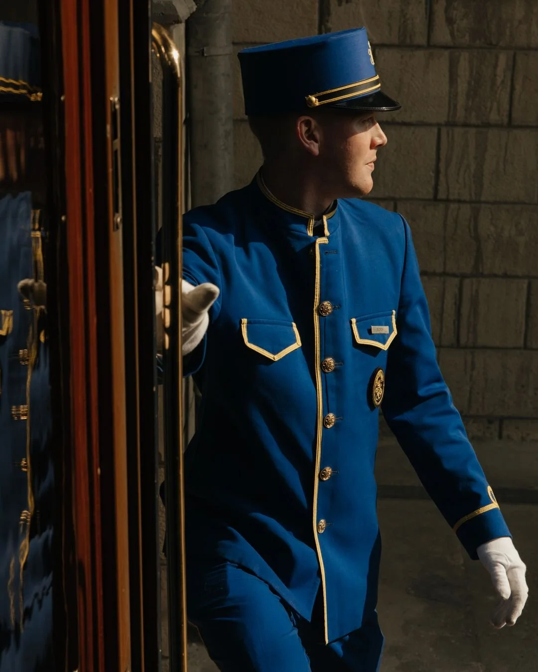 A uniformed bellhop in a blue suit with gold trim and buttons, also wearing a matching blue hat with gold accents, standing at an entrance with a luggage cart.